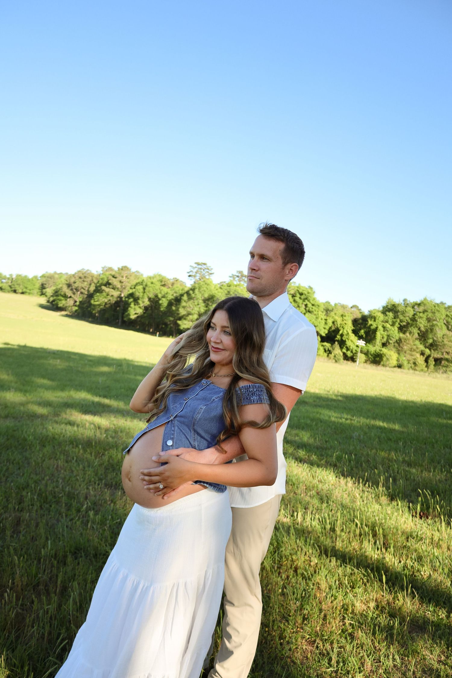 A couple poses outdoors in a green field, the man embracing a pregnant woman in a white skirt and blue top.
