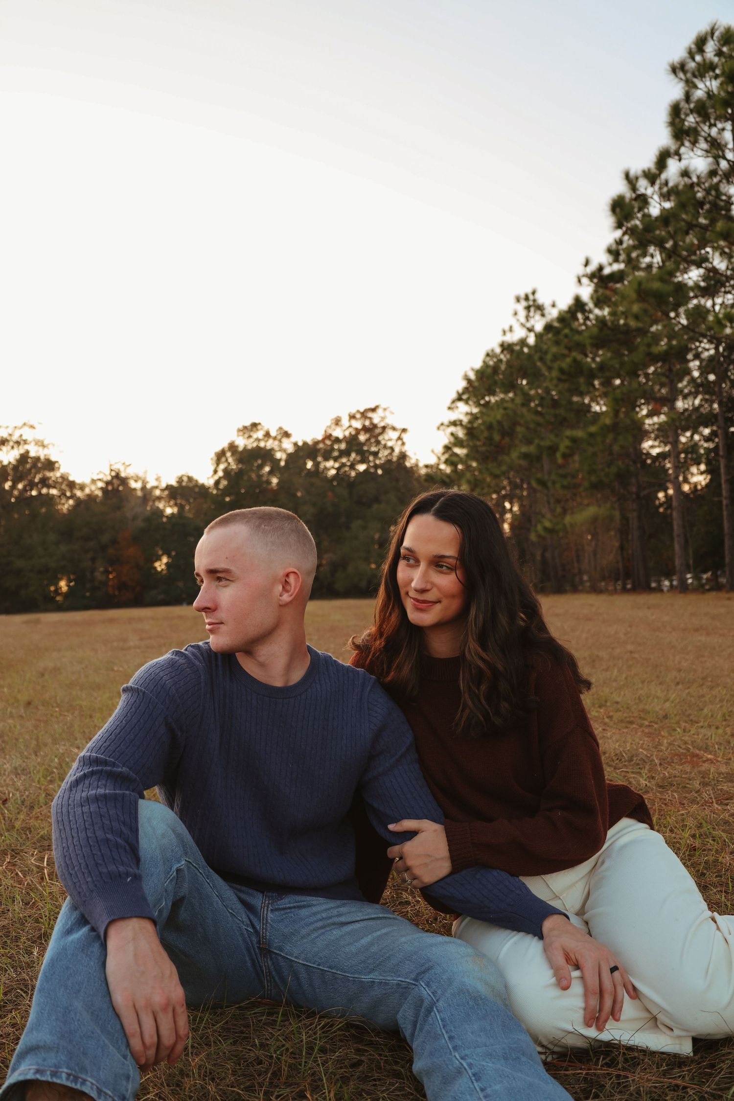 Couple sitting together on grass at sunset in autumn with pine trees in background.