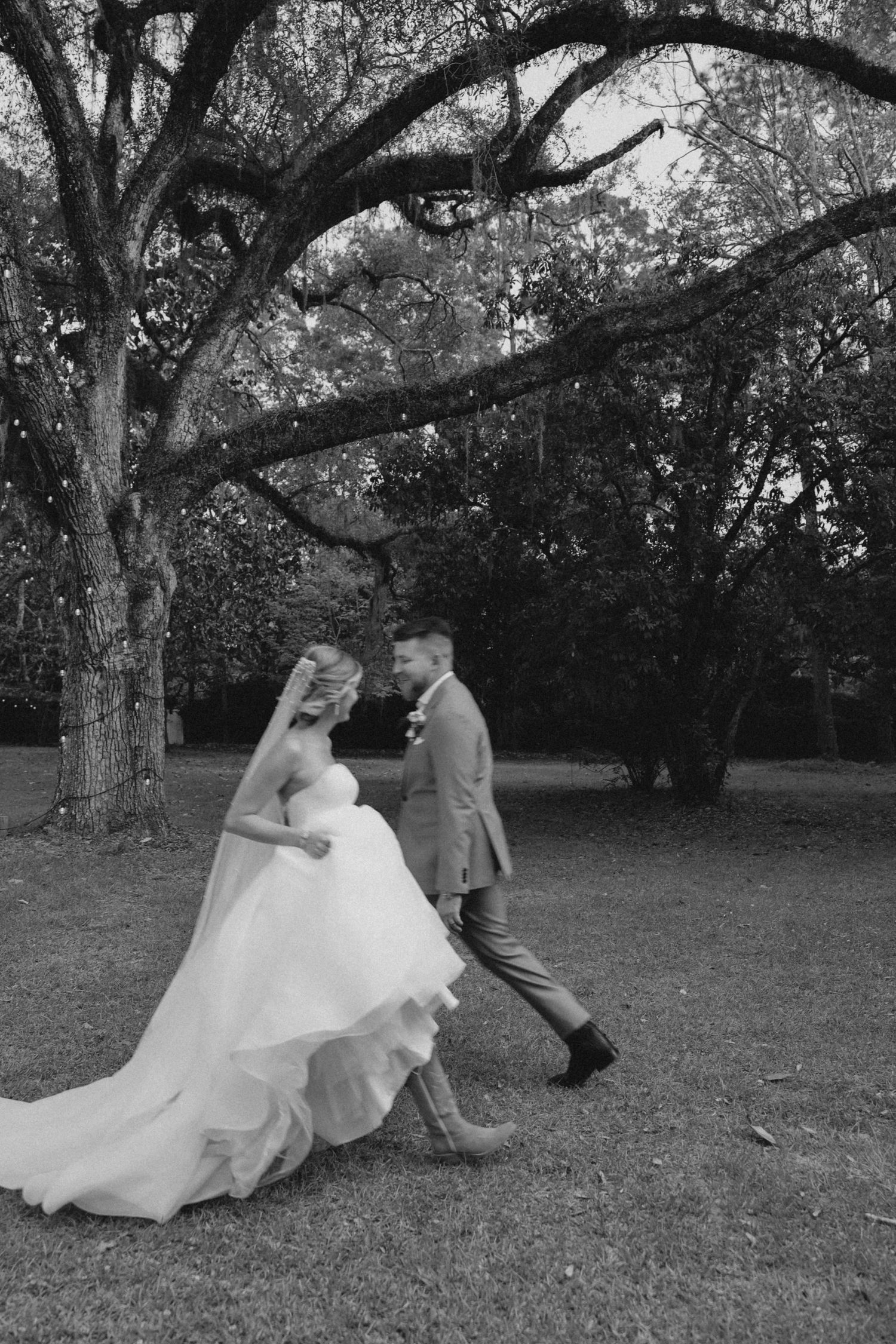 Black and white photo of a bride and groom walking hand in hand under a large oak tree on their wedding day.