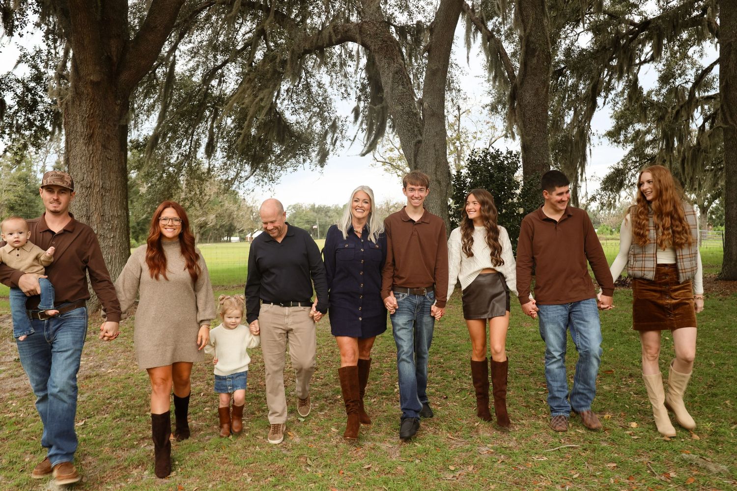 Large family of adults and children walking hand in hand across a grassy lawn beneath oak trees draped in Spanish moss.