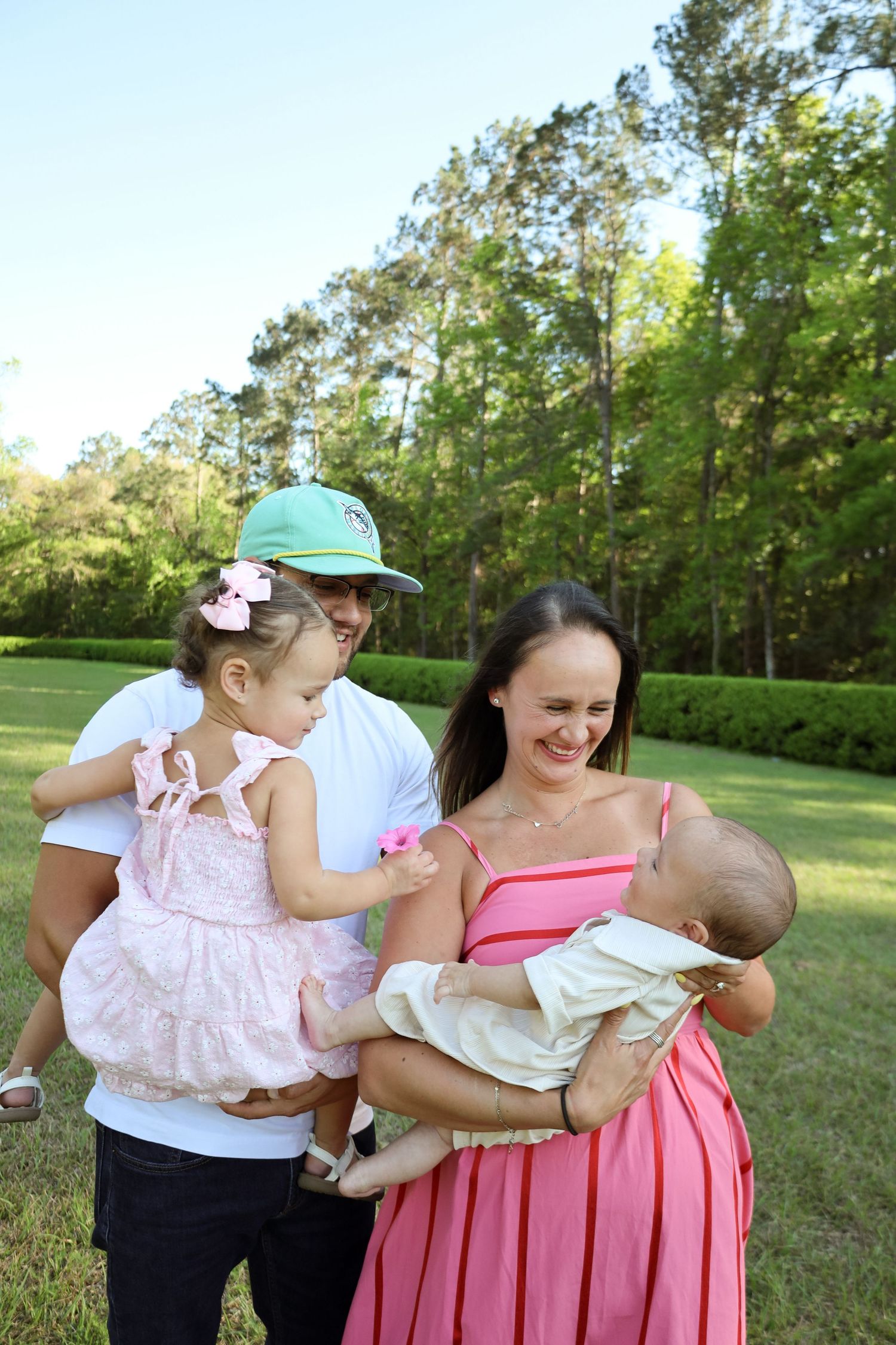 A family wearing pink and white outfits poses together in a grassy outdoor setting with trees in the background.