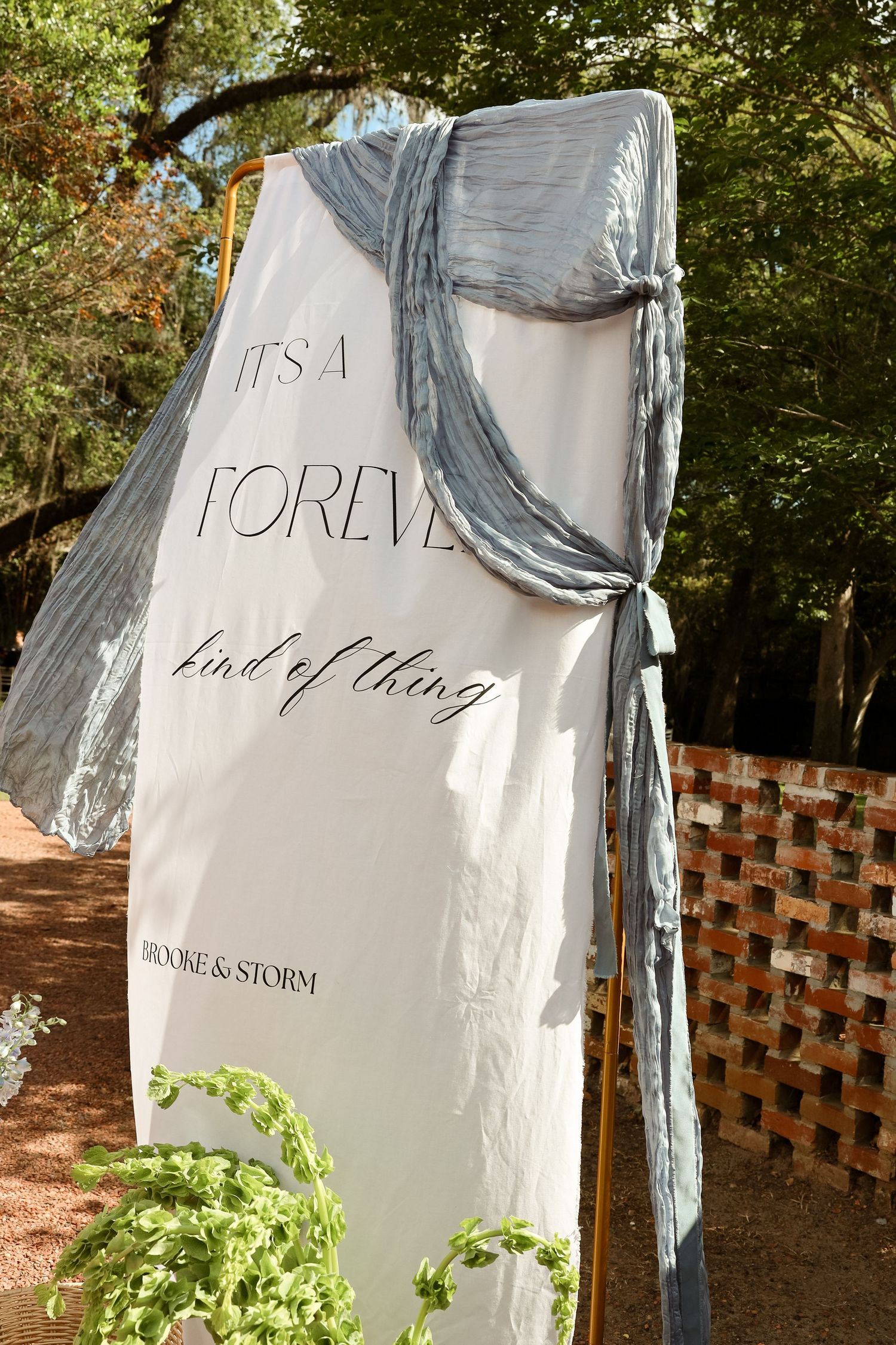 Outdoor wedding sign reading It's A Forever Kind of Thing for Brooke & Storm, draped with blue fabric outdoors.