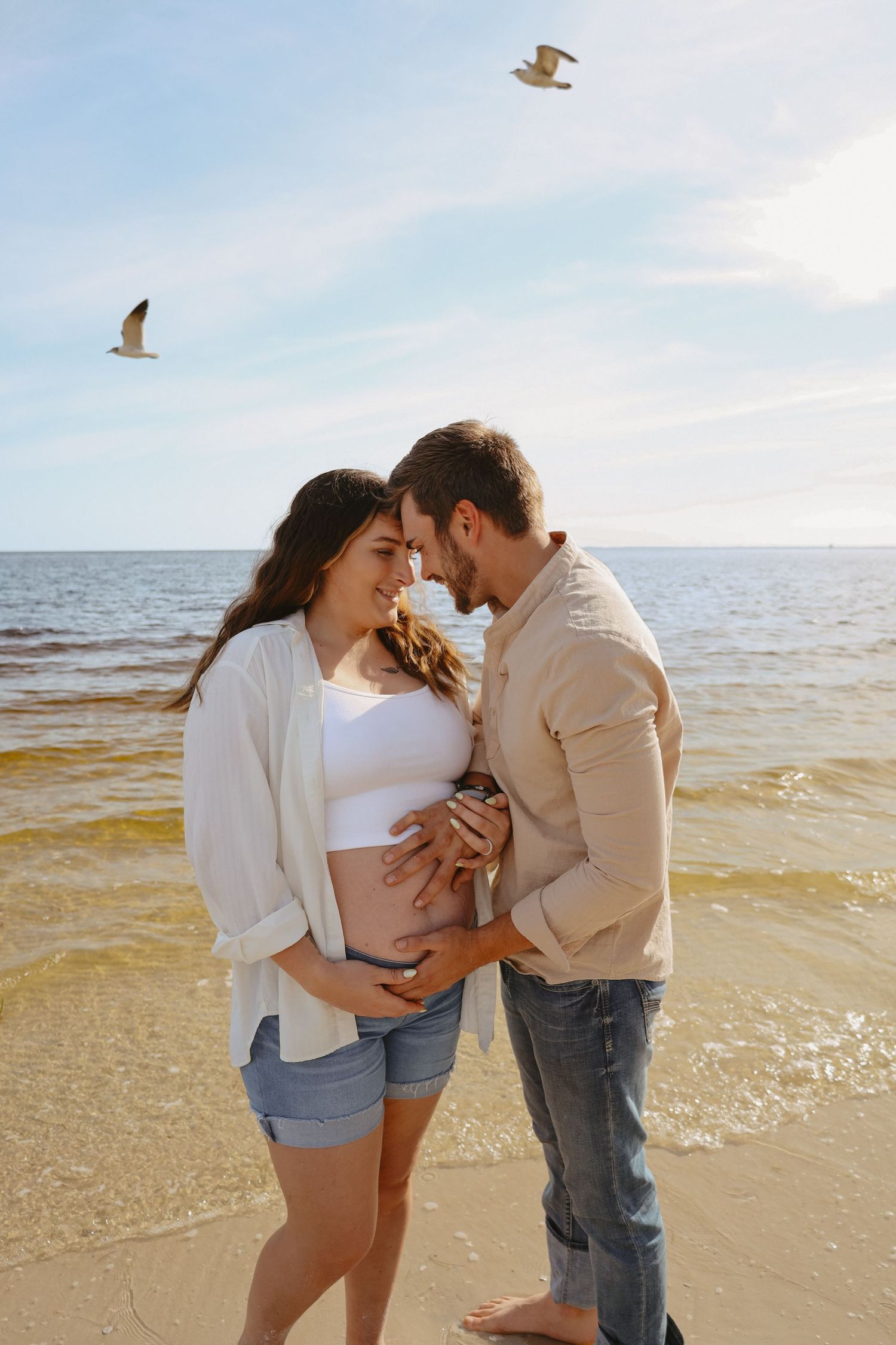 A couple shares an intimate moment at the beach during sunset, showcasing a pregnancy photoshoot.