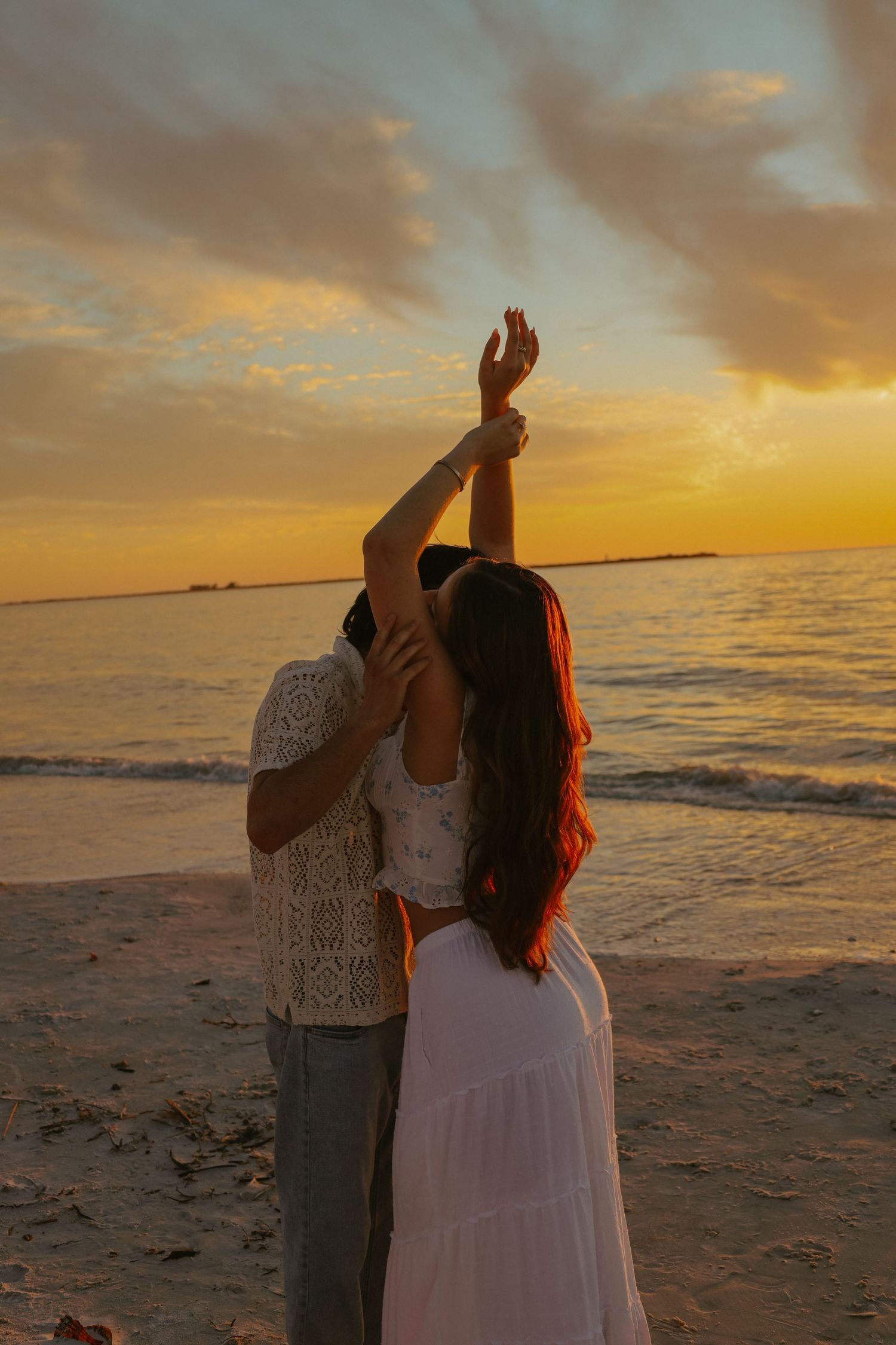 A couple embraces on a sandy beach at sunset with golden clouds and ocean waves in the background.