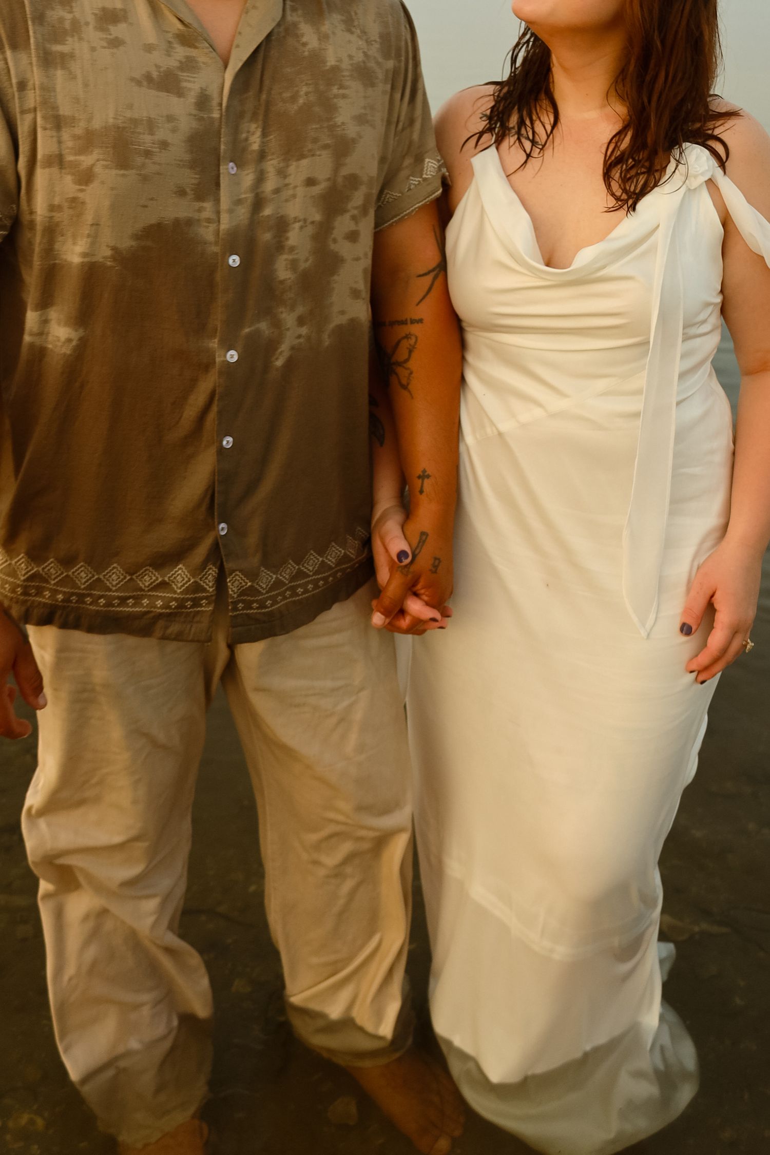 Bride in white cowl-neck gown holds hands with groom in brown patterned shirt on a beach.