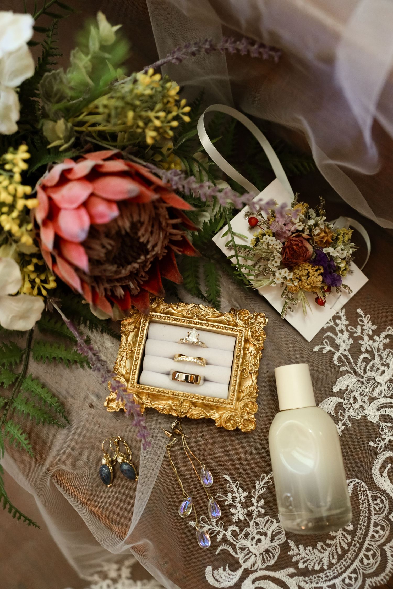 Wedding flat lay with gold ring box, engagement rings, floral bouquet, perfume bottle, earrings, and lace veil on wooden surface.