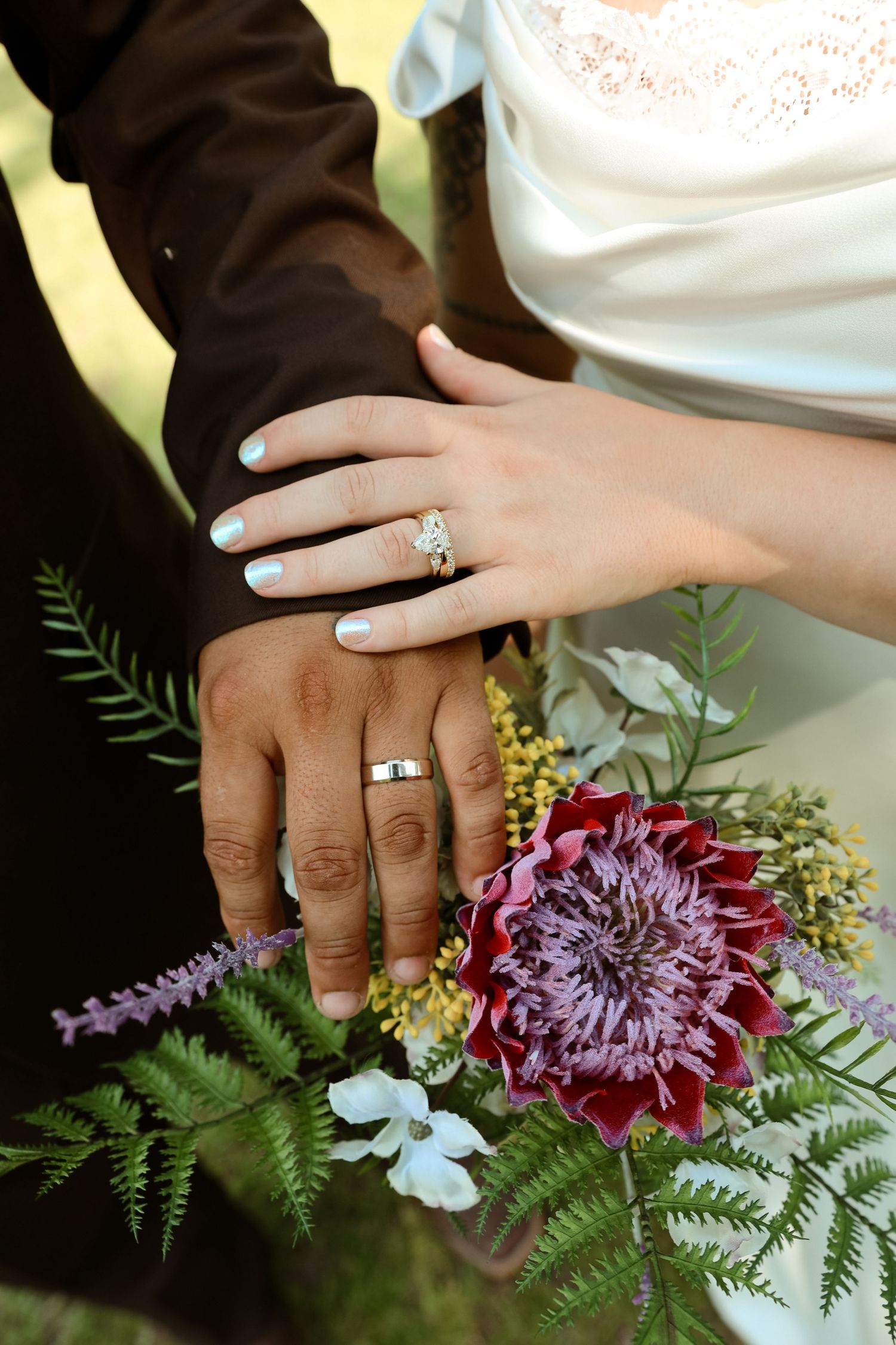 Newlywed couple displaying wedding rings on hands resting over a bridal bouquet with purple dahlia and ferns.