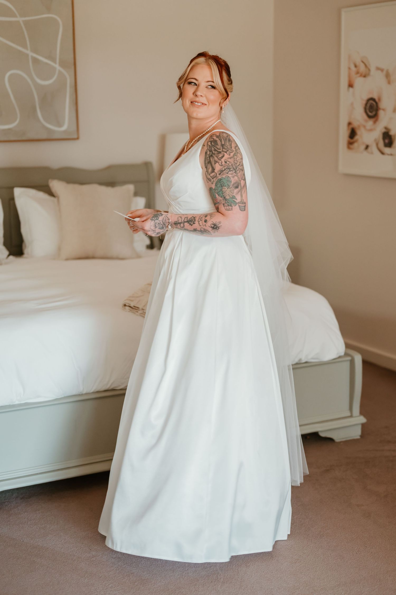 A bride in a classic white wedding gown stands in an elegantly decorated bedroom getting ready for her ceremony.