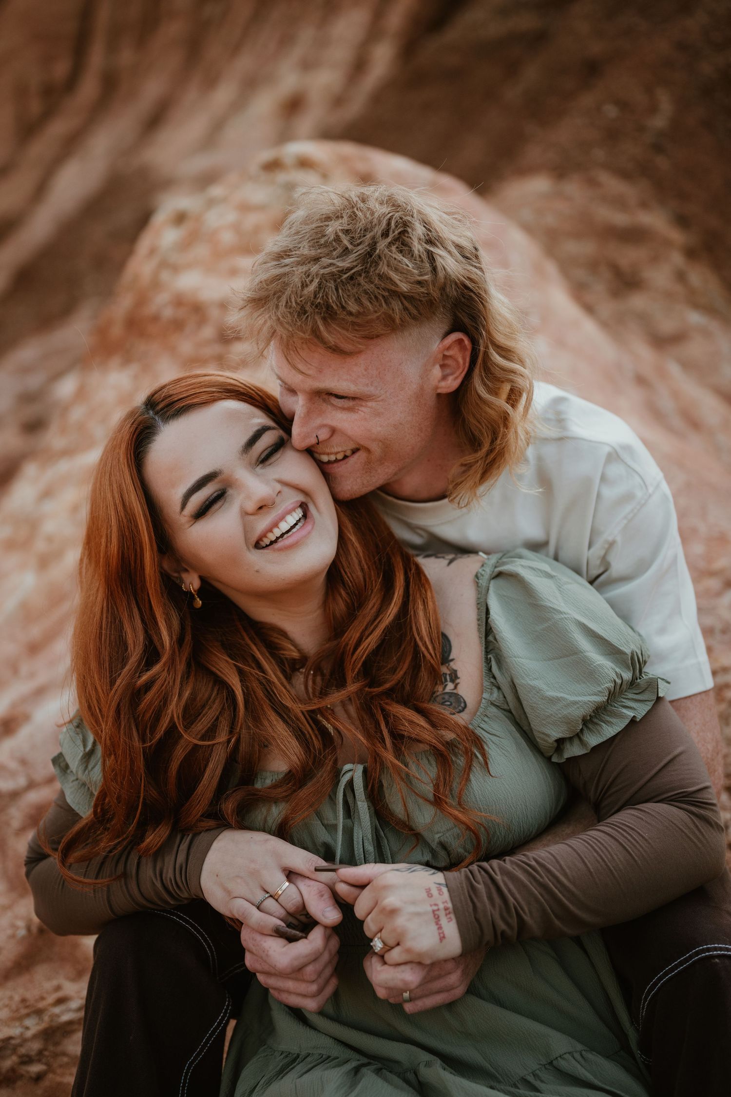 A joyful couple share an intimate moment against a rusty orange rock formation in warm natural lighting.