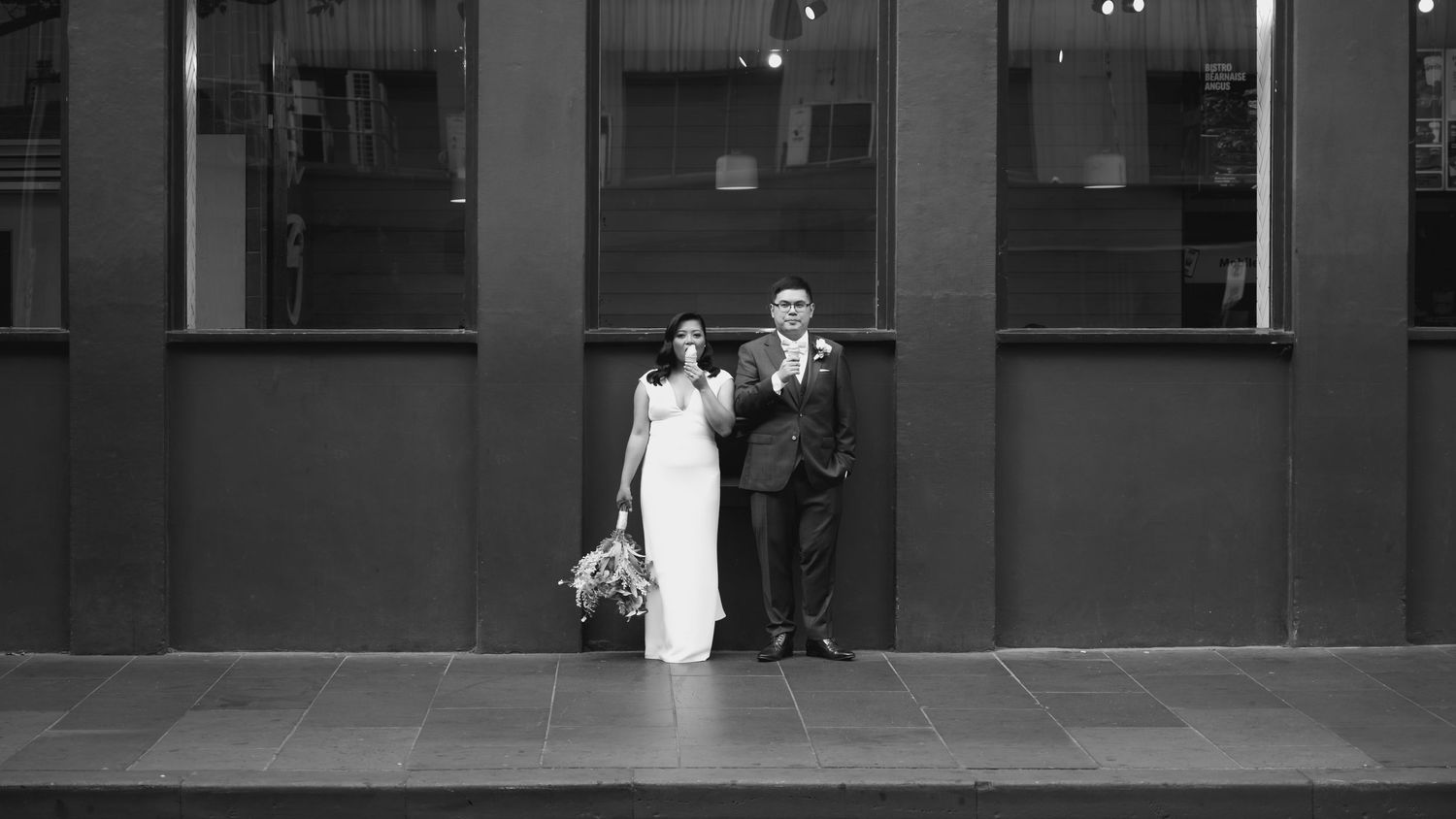 A black and white panoramic wedding photo sequence showing the couple walking across a modern building exterior.