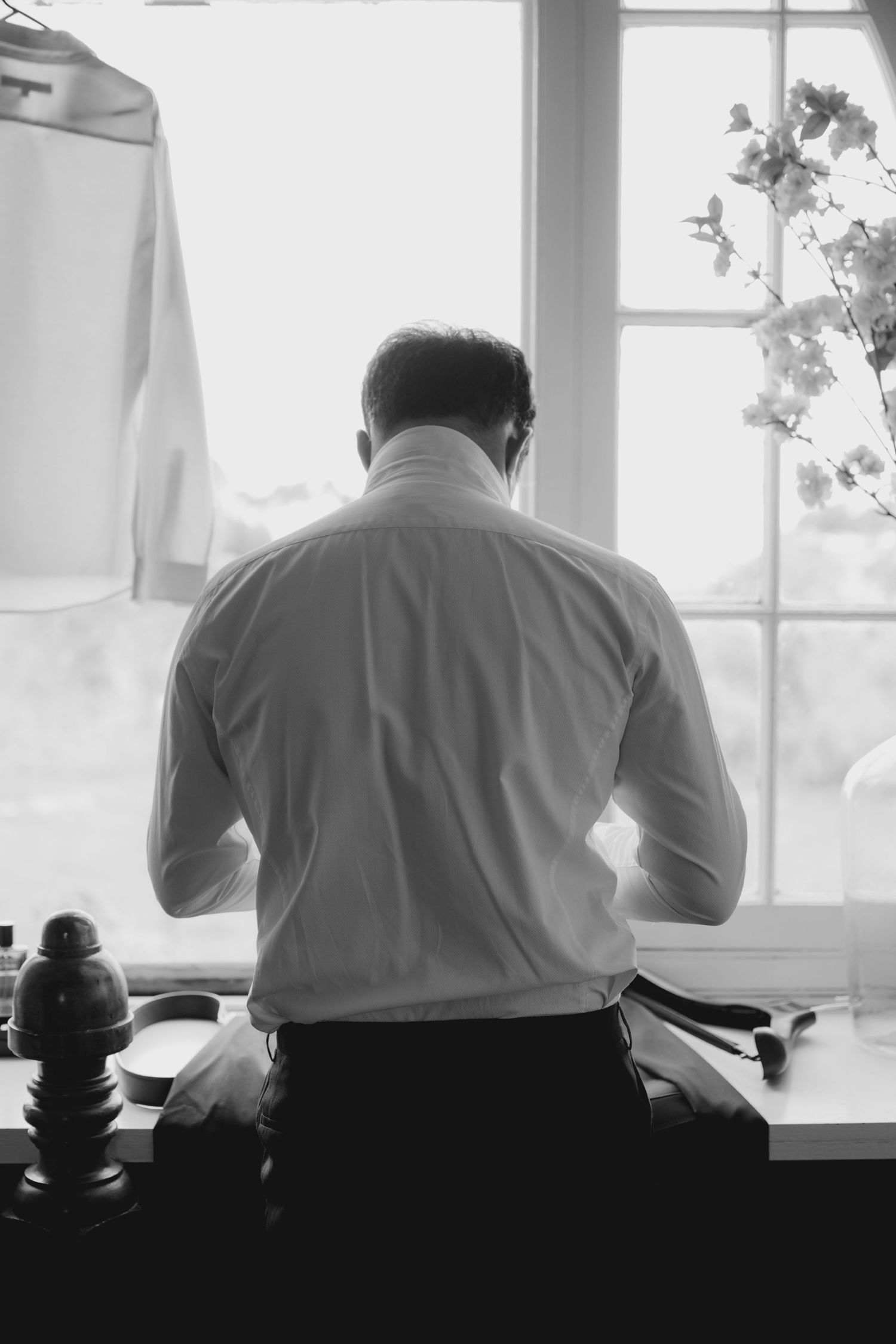 A person in a white dress shirt stands at a window, viewed from behind in black and white photography.