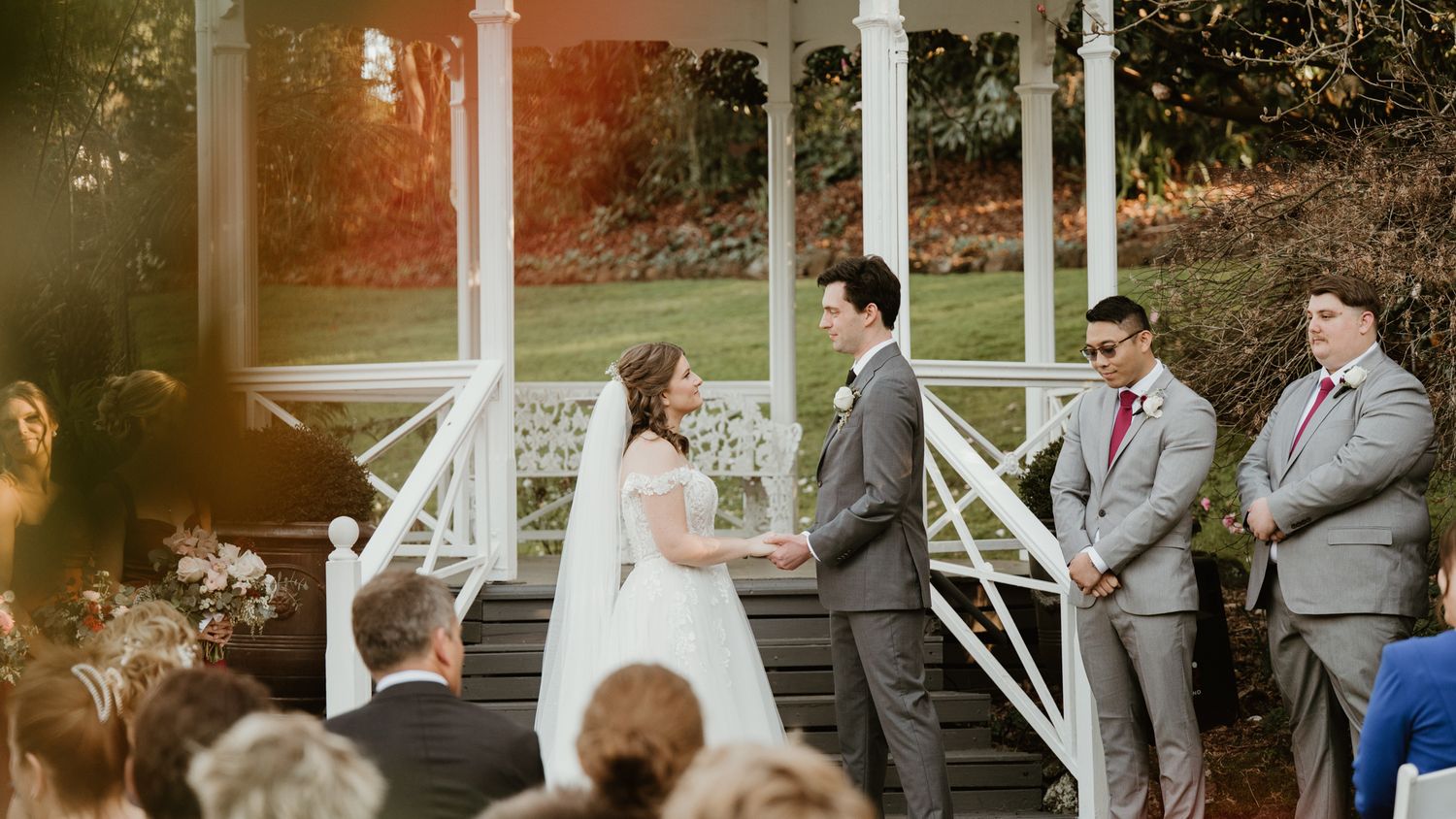 Bride and groom exchange vows at an outdoor wedding ceremony beneath a white gazebo, with groomsmen standing nearby.