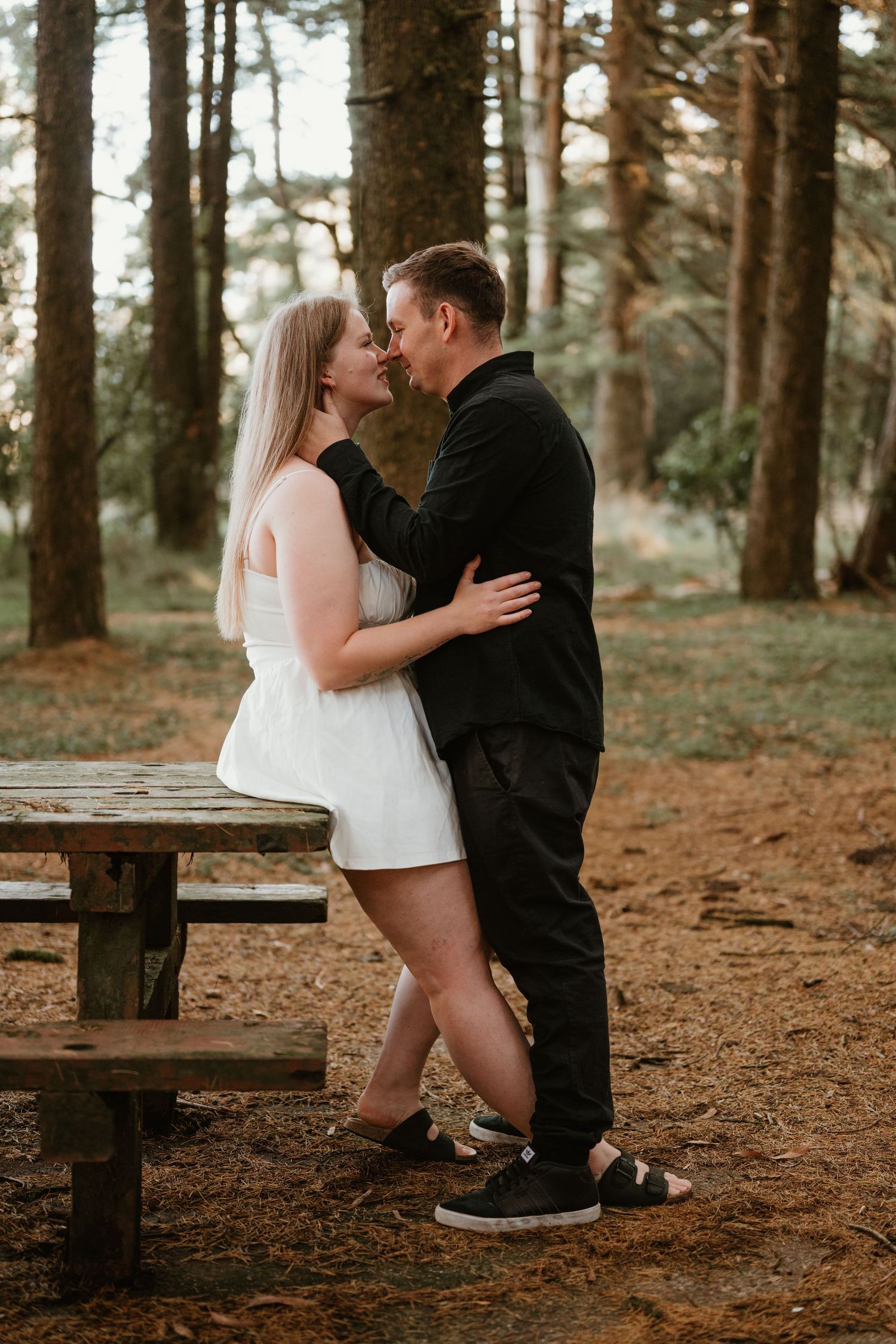 Two figures share an intimate moment on a wooden bench in a wooded forest setting.