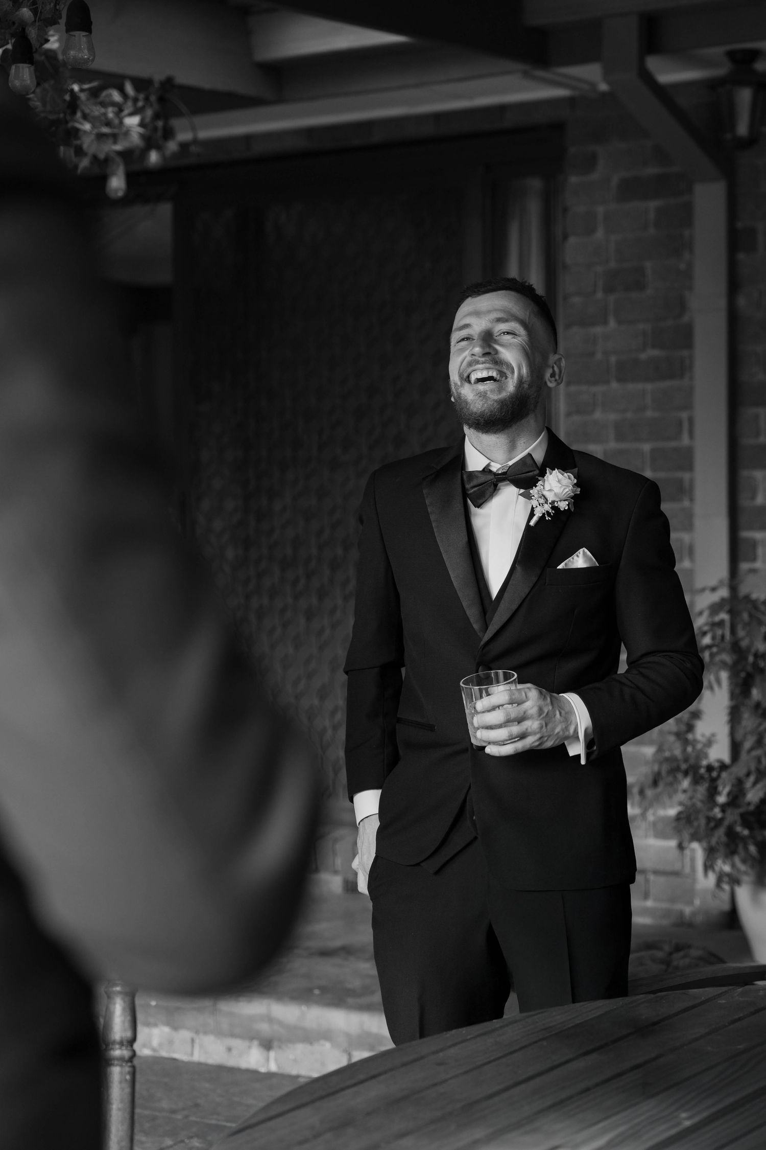 Person in formal black suit standing outside brick building holding drink and laughing.