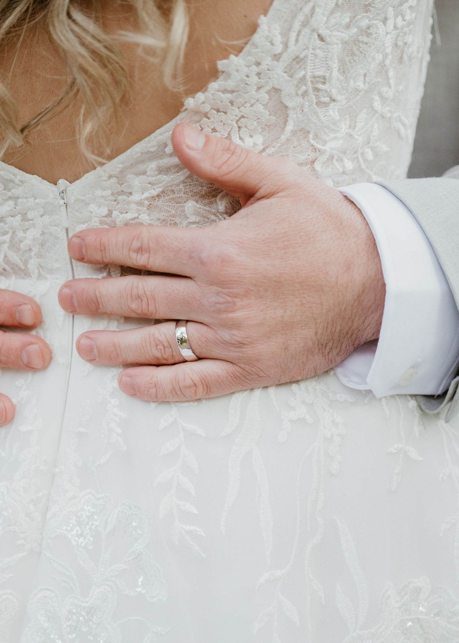 Close-up of groom's hand with wedding band resting on bride's white lace wedding dress.