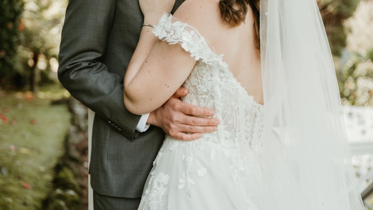 Bride and groom embracing outdoors, showcasing lace wedding dress with off-shoulder detail and groom in gray suit.
