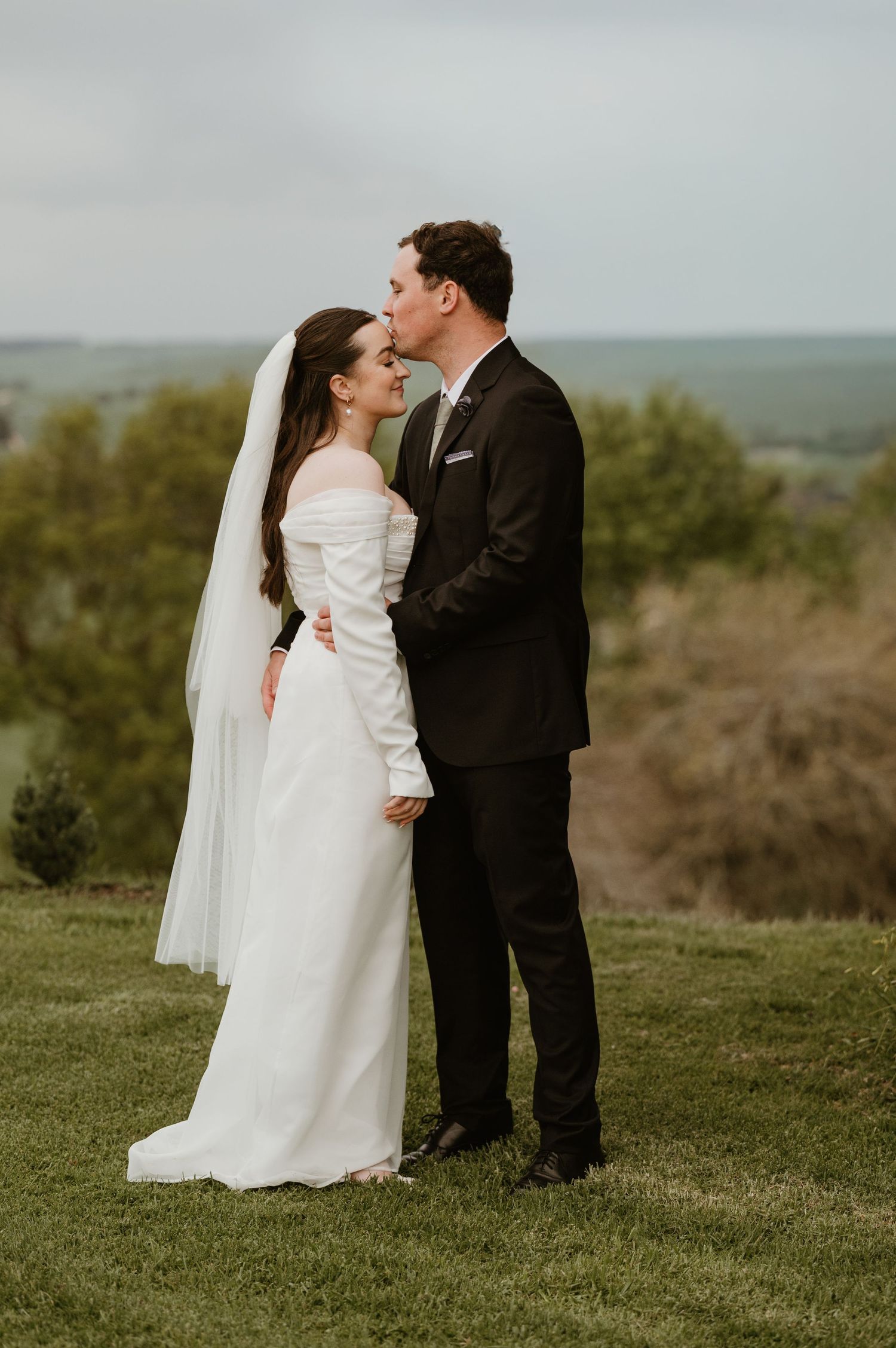 A romantic wedding photo of a couple embracing on a hilltop with a scenic countryside view in the background.