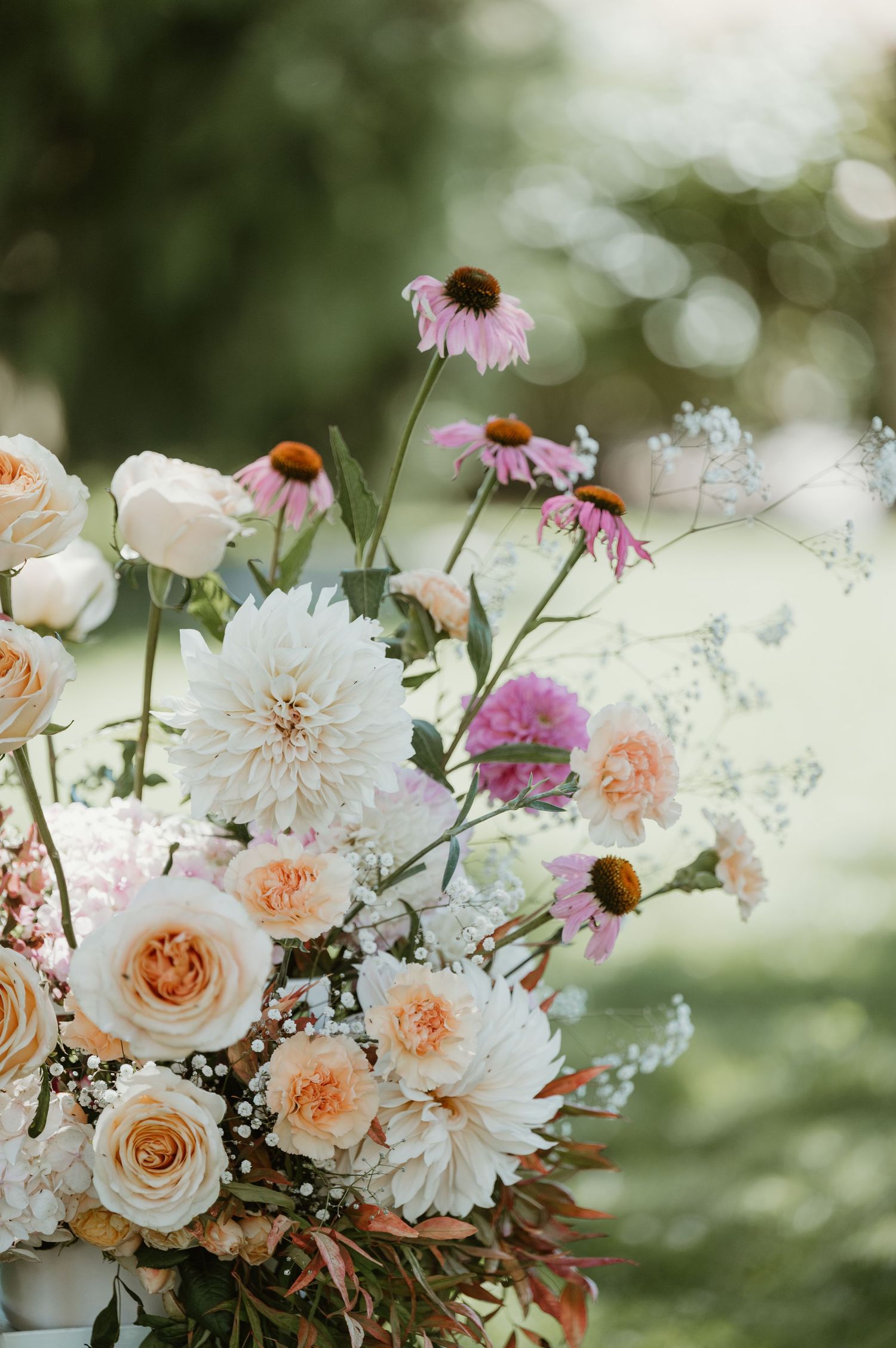 A romantic garden-inspired floral arrangement with peach roses, white dahlias, and purple coneflowers against a blurred background.