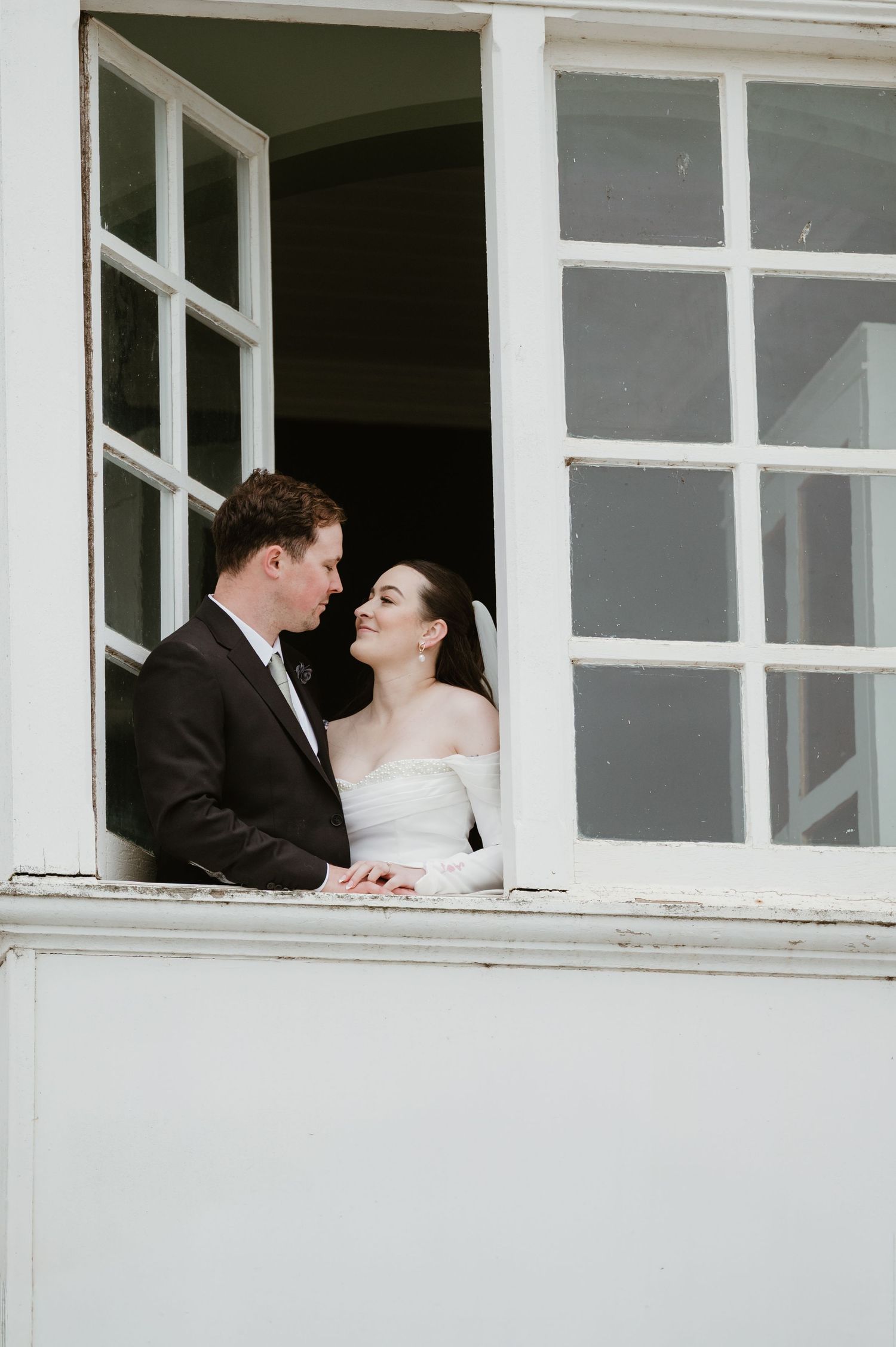 A romantic wedding couple shares a tender moment through a white-framed window.