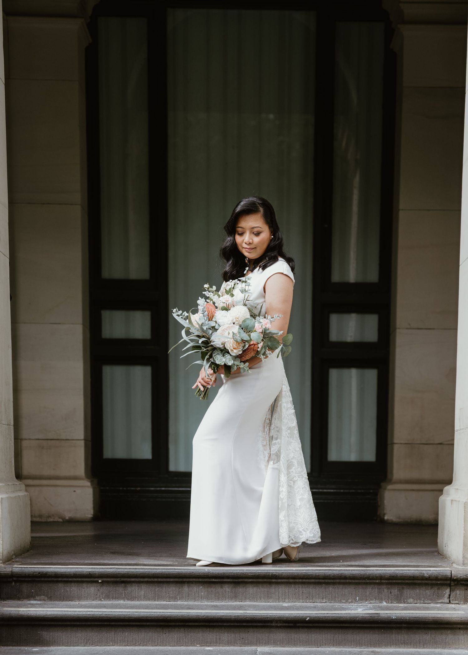 Bride in a white wedding dress holds a bouquet while standing on a covered porch with white columns.