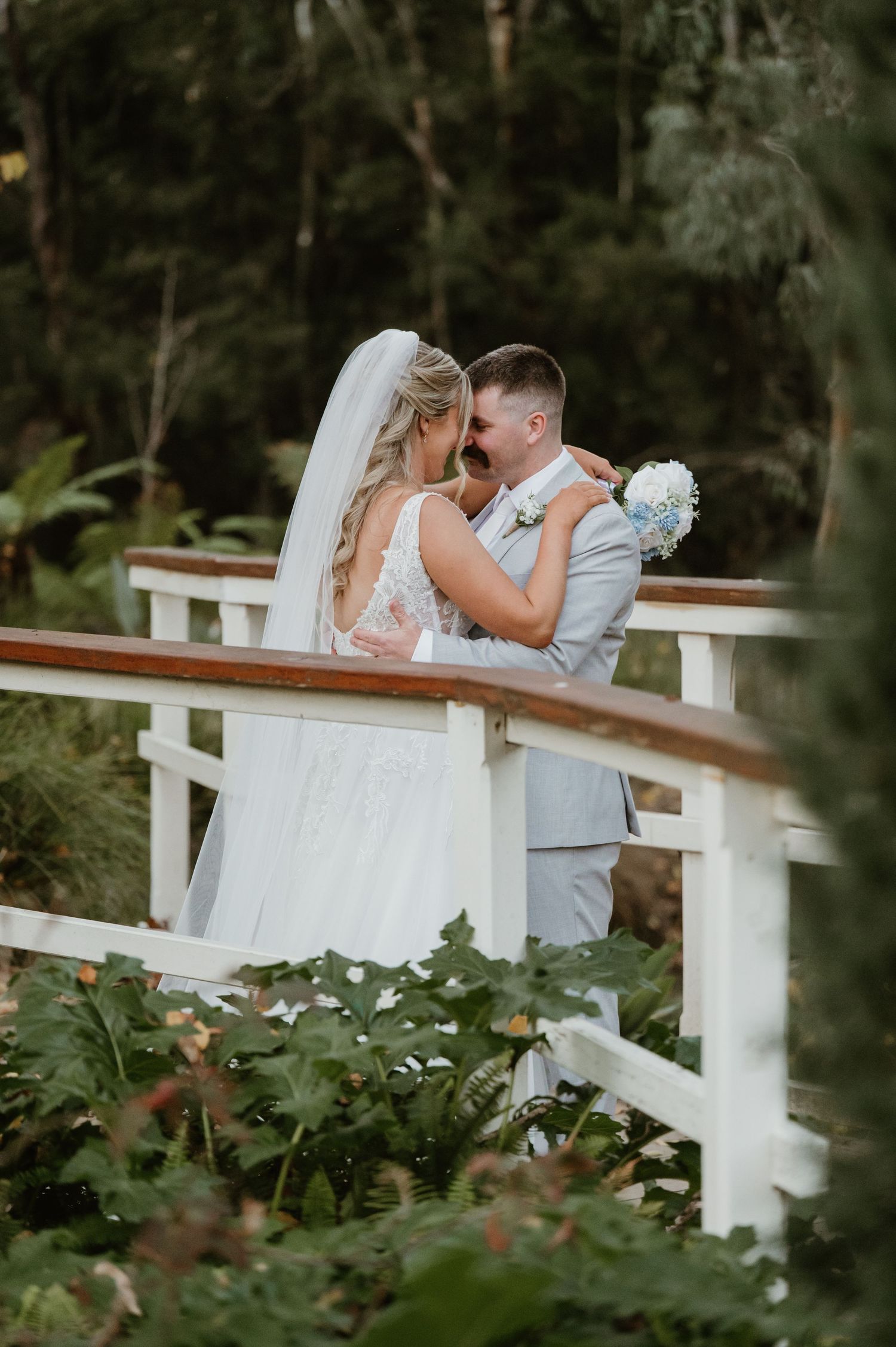 Bride and groom share a romantic moment on a white wooden bridge surrounded by lush greenery on their wedding day.