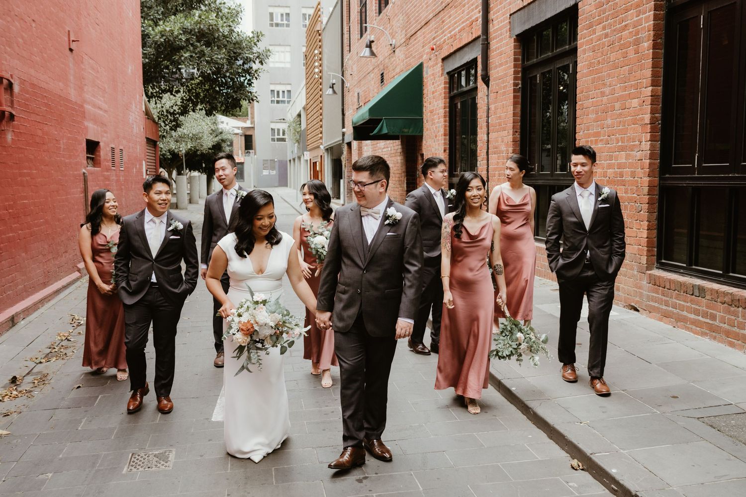A wedding party walks down an urban street with brick buildings, wearing formal attire and carrying bouquets.
