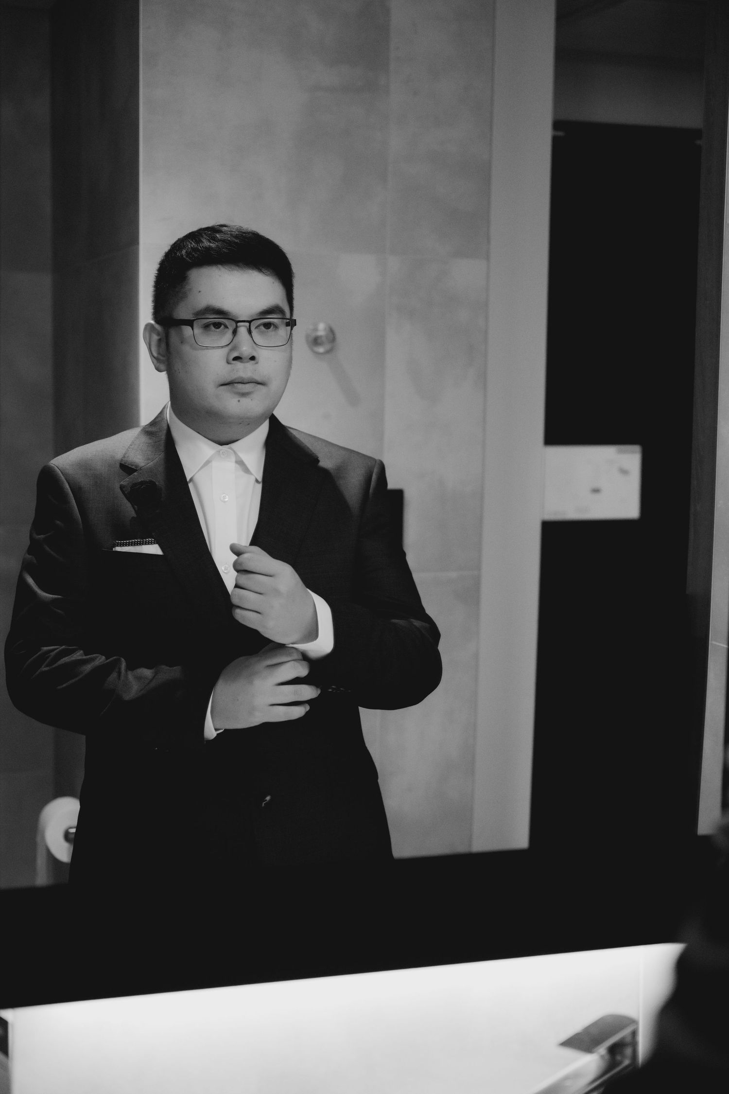 Black and white portrait of someone in a formal suit and glasses adjusting their cuff in an elegant indoor setting.