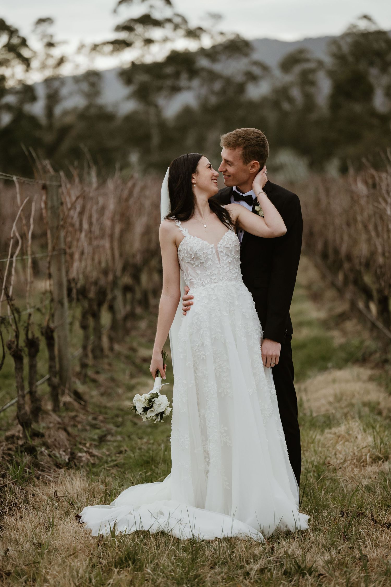 A couple poses intimately in a vineyard during sunset with the bride wearing a flowing white lace gown.