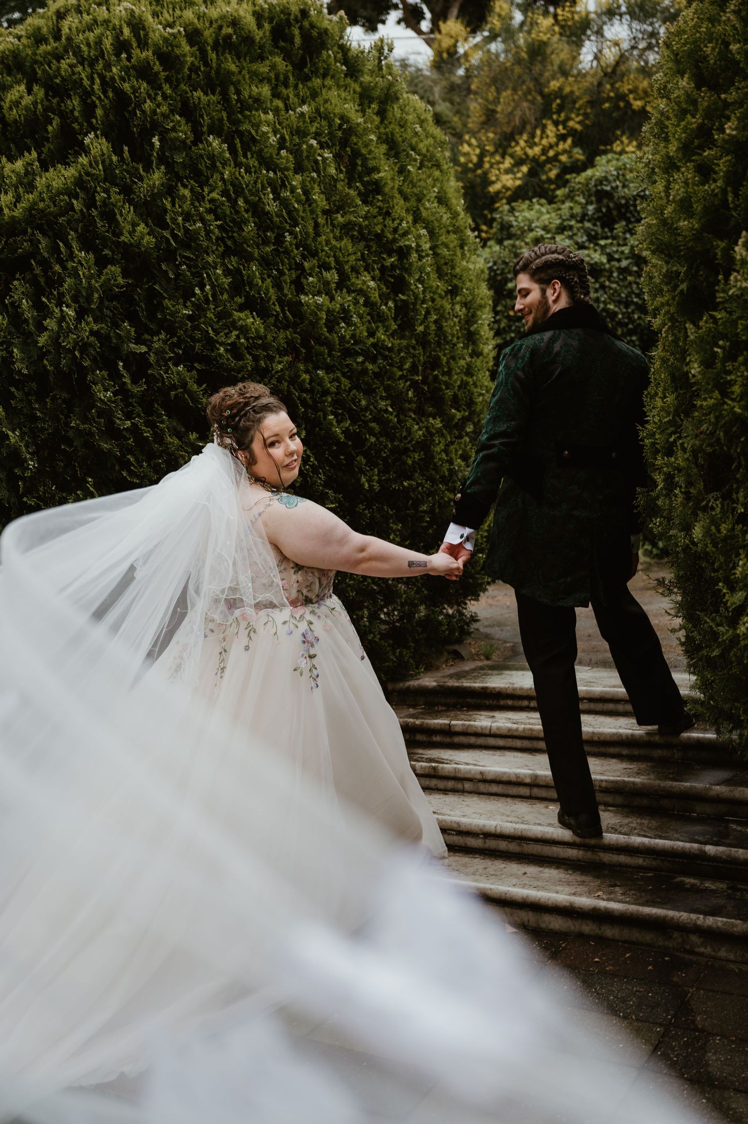 A couple in wedding attire playfully pose on garden steps with flowing white dress and veil creating dramatic movement.