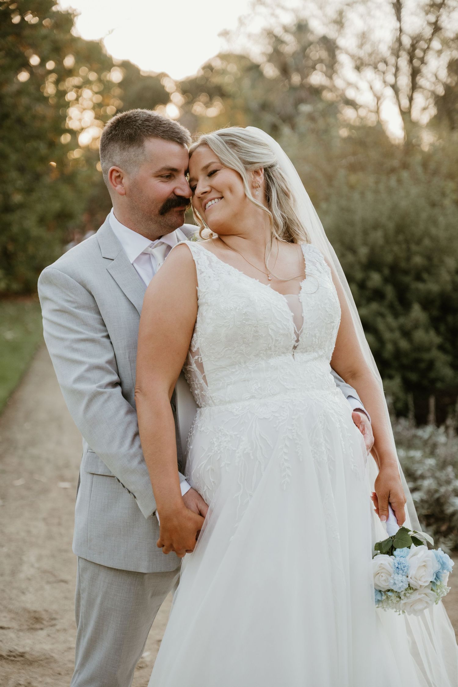 A bride and groom share a romantic moment outdoors, she in a lace gown holding a blue bouquet, he in a gray suit.