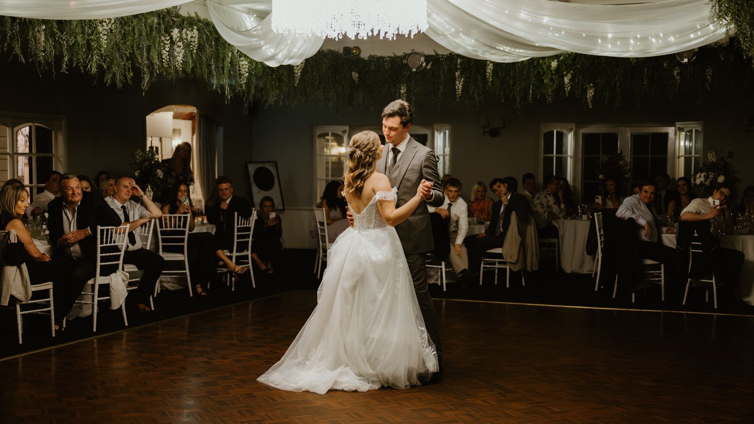 Bride and groom share first dance at elegant wedding reception with greenery ceiling decor and guests seated around.