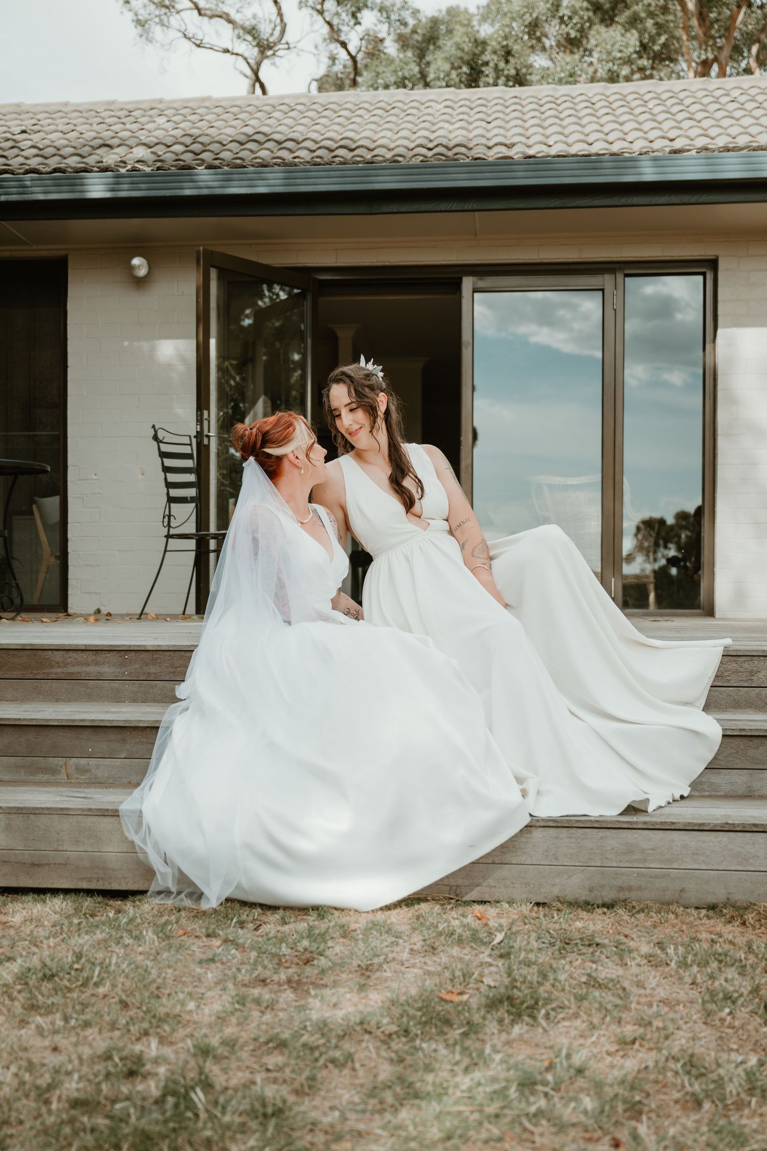 Two people in white wedding dresses share a romantic moment on an outdoor patio.