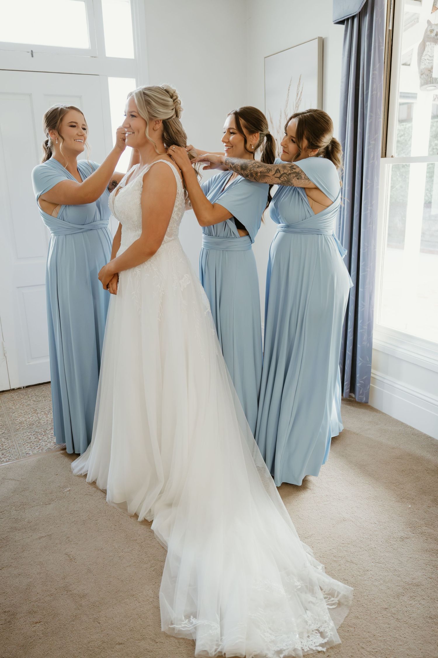 Bride in white gown assisted by three bridesmaids in light blue dresses during wedding preparations.
