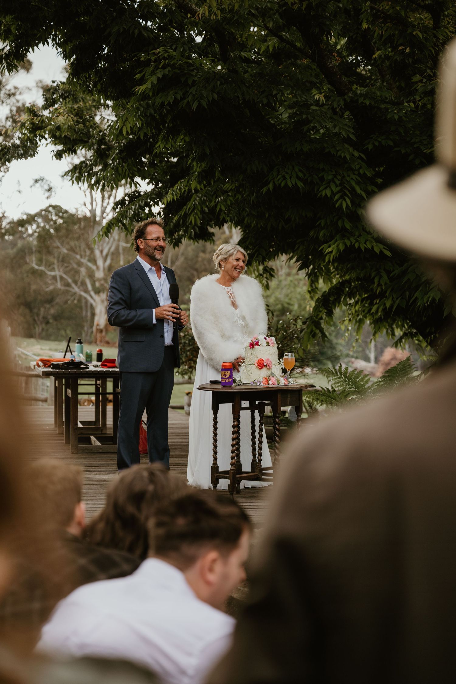 Wedding guests gather outdoors under trees at a rustic reception venue with wooden picnic tables.