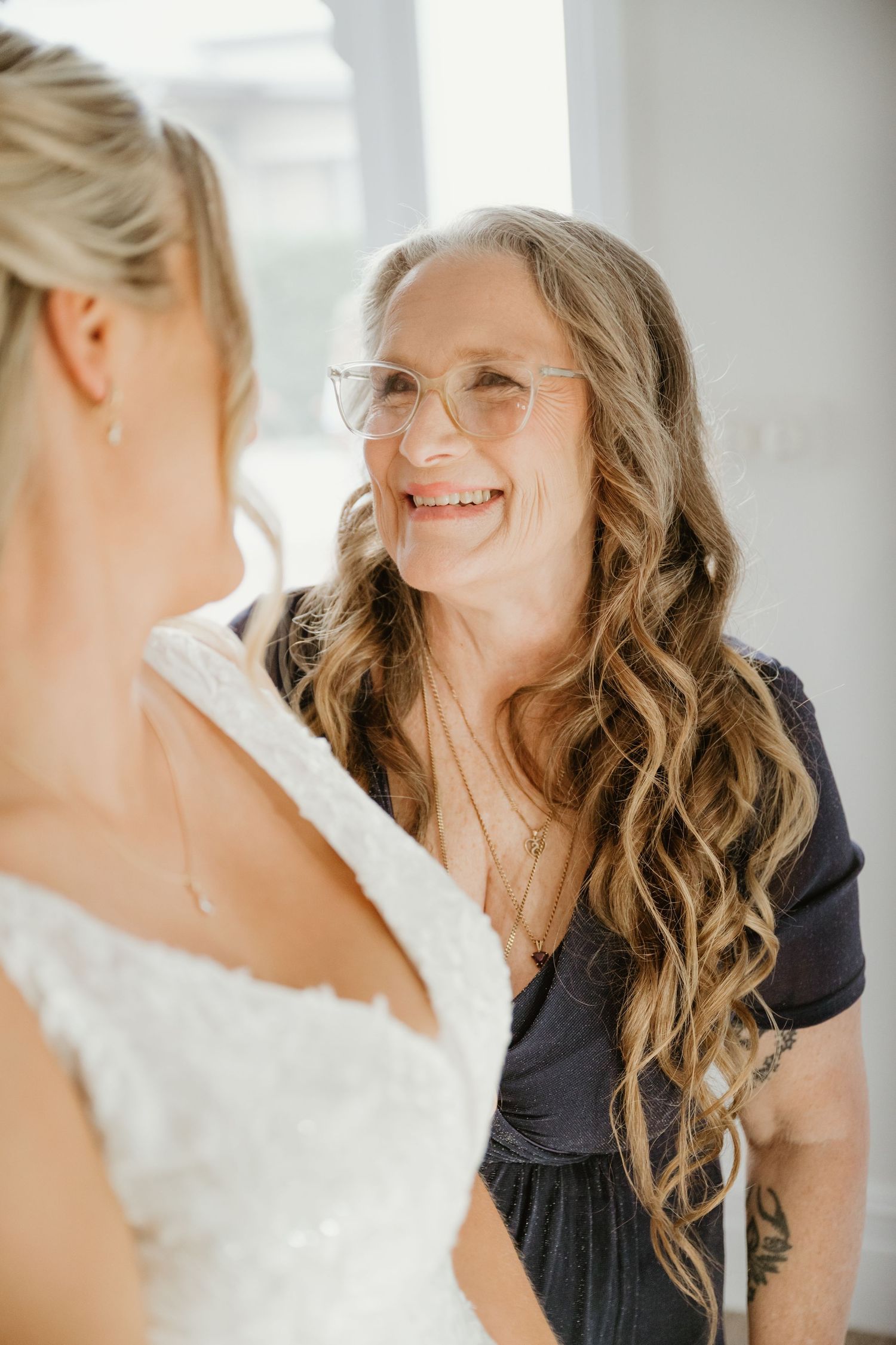 A smiling older woman with curly hair and glasses looks warmly at a bride in a white lace wedding dress.