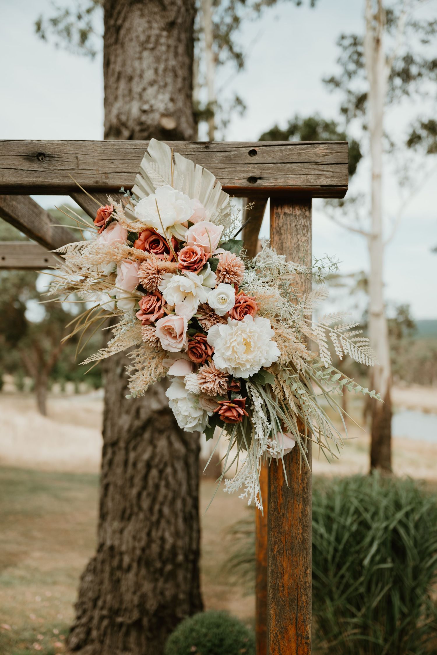Rustic wedding arch floral arrangement with pink roses and dried pampas grass against a woodland backdrop.