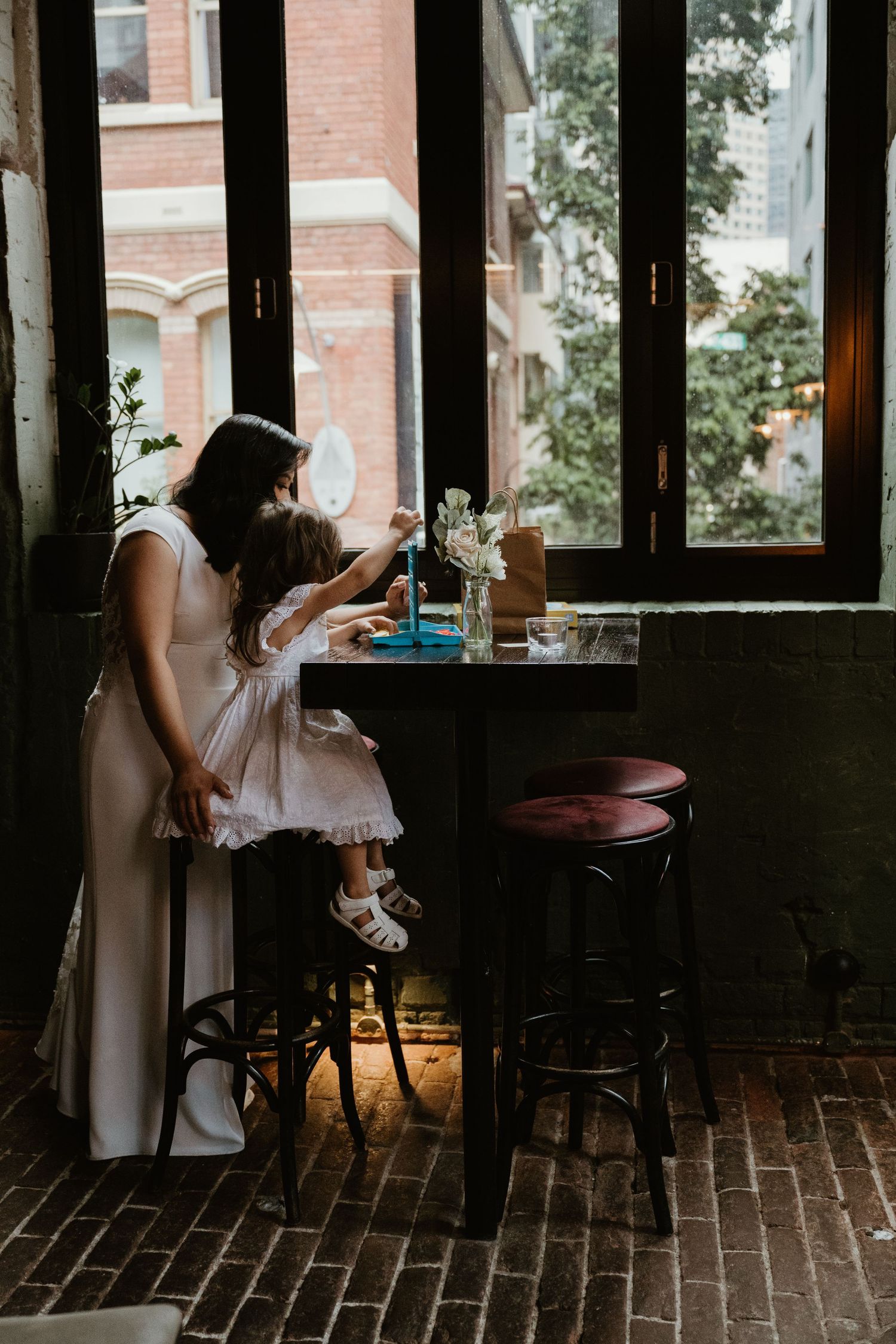 Two people share a tender moment at a cozy cafe table by a window with exposed brick walls and vintage decor.