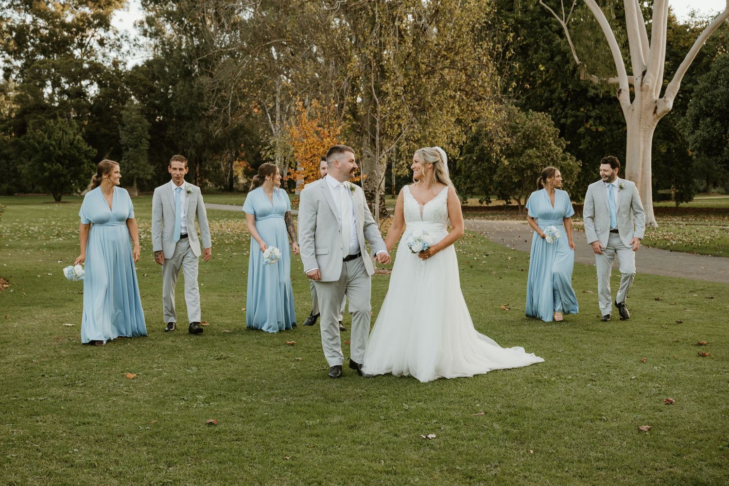 Wedding party walks on green lawn, bride in white gown with groom, bridesmaids in blue dresses and groomsmen in gray suits.