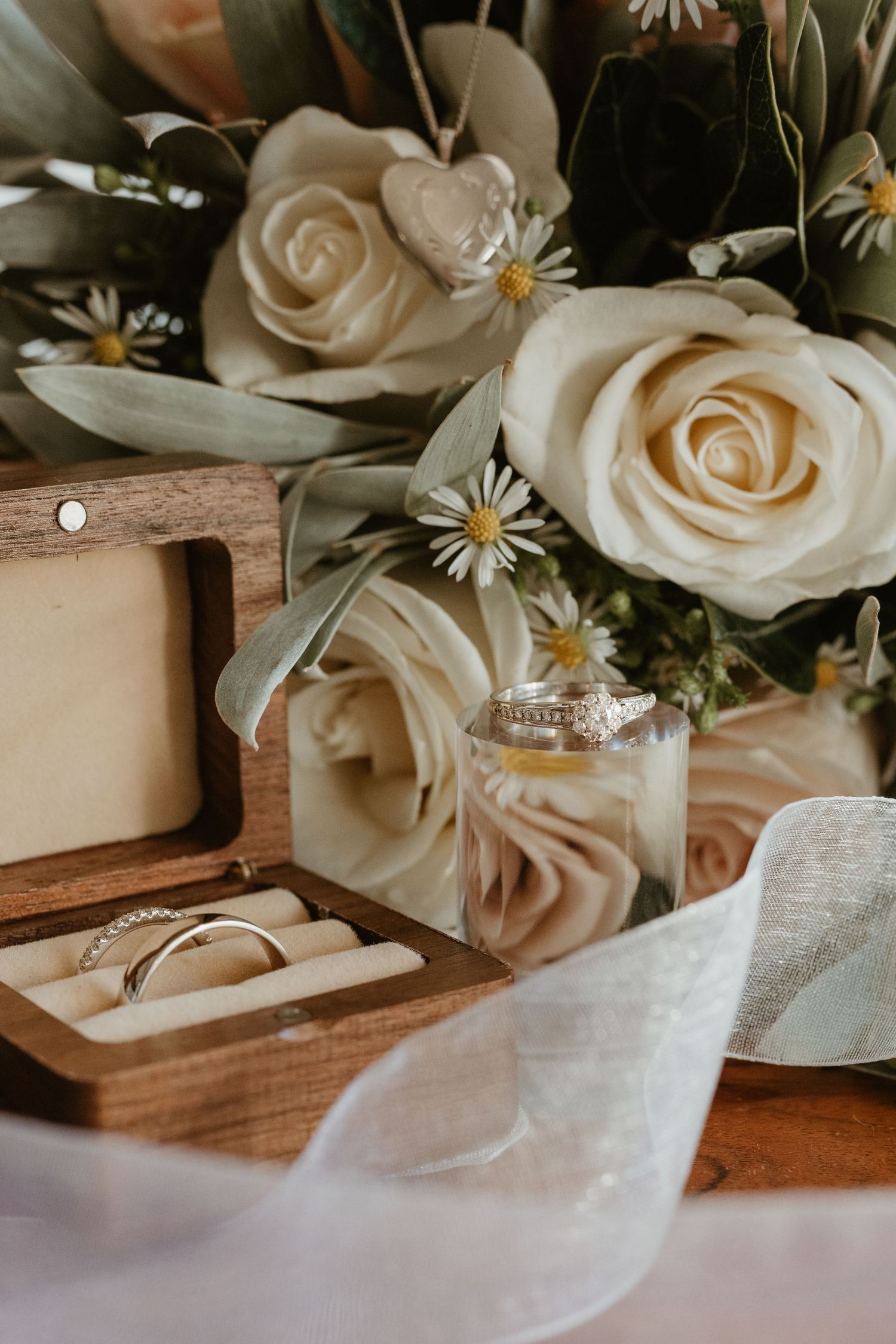 Wedding details including white roses, ribbon, and rings displayed on a rustic wooden surface.