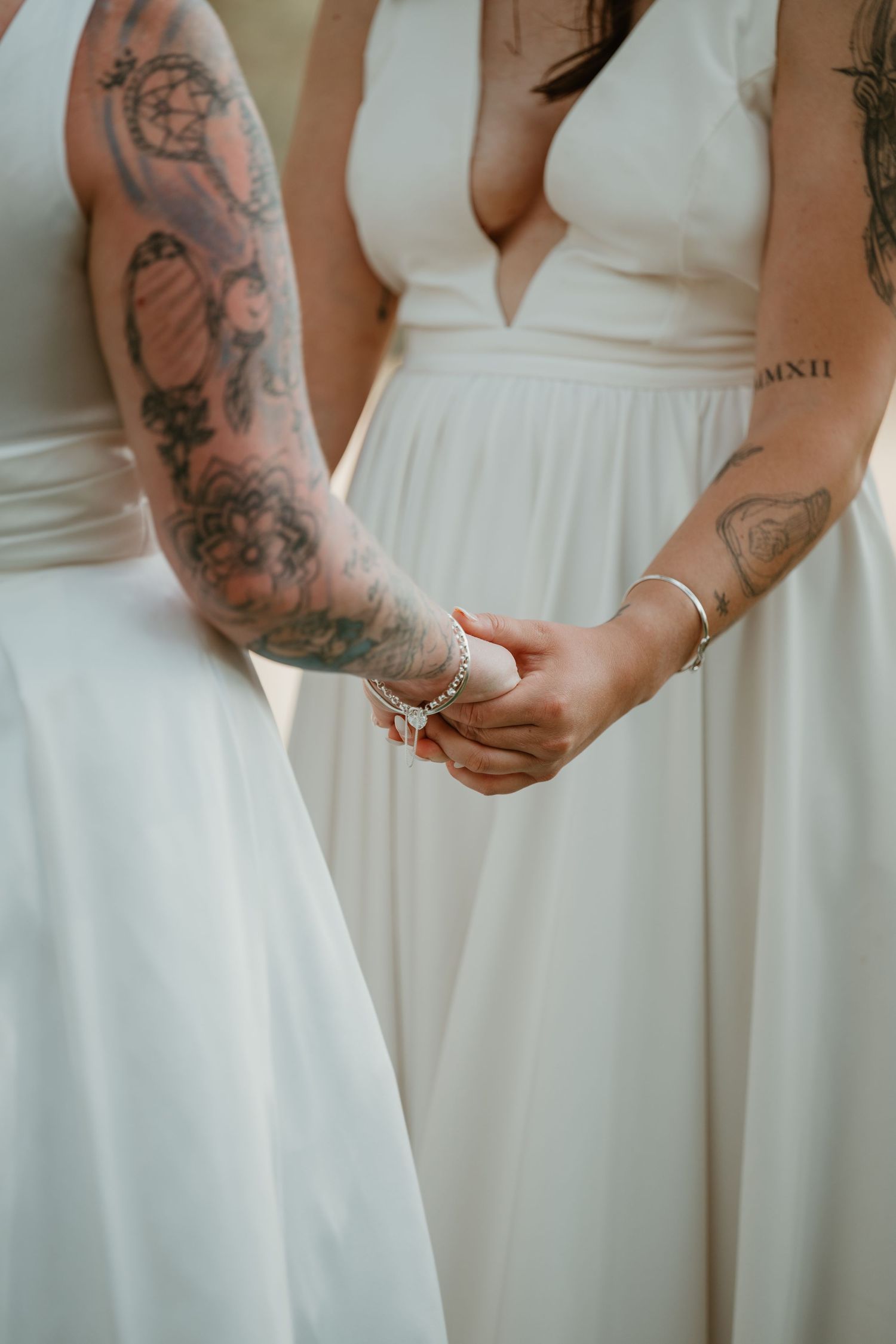 Close-up of two people in white dresses holding hands, showing detailed arm tattoos.