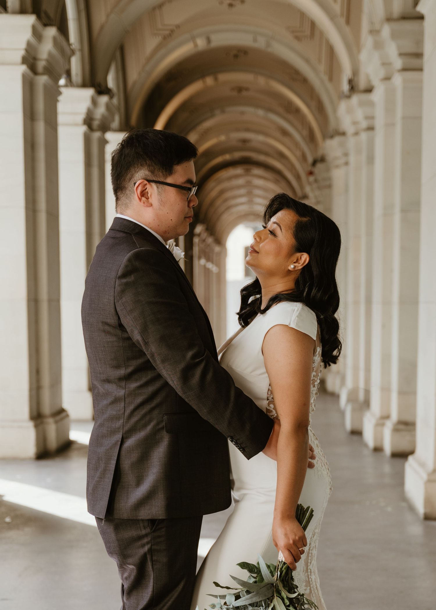 A couple shares an intimate moment in a white arched colonnade hallway while dressed in formal wedding attire.