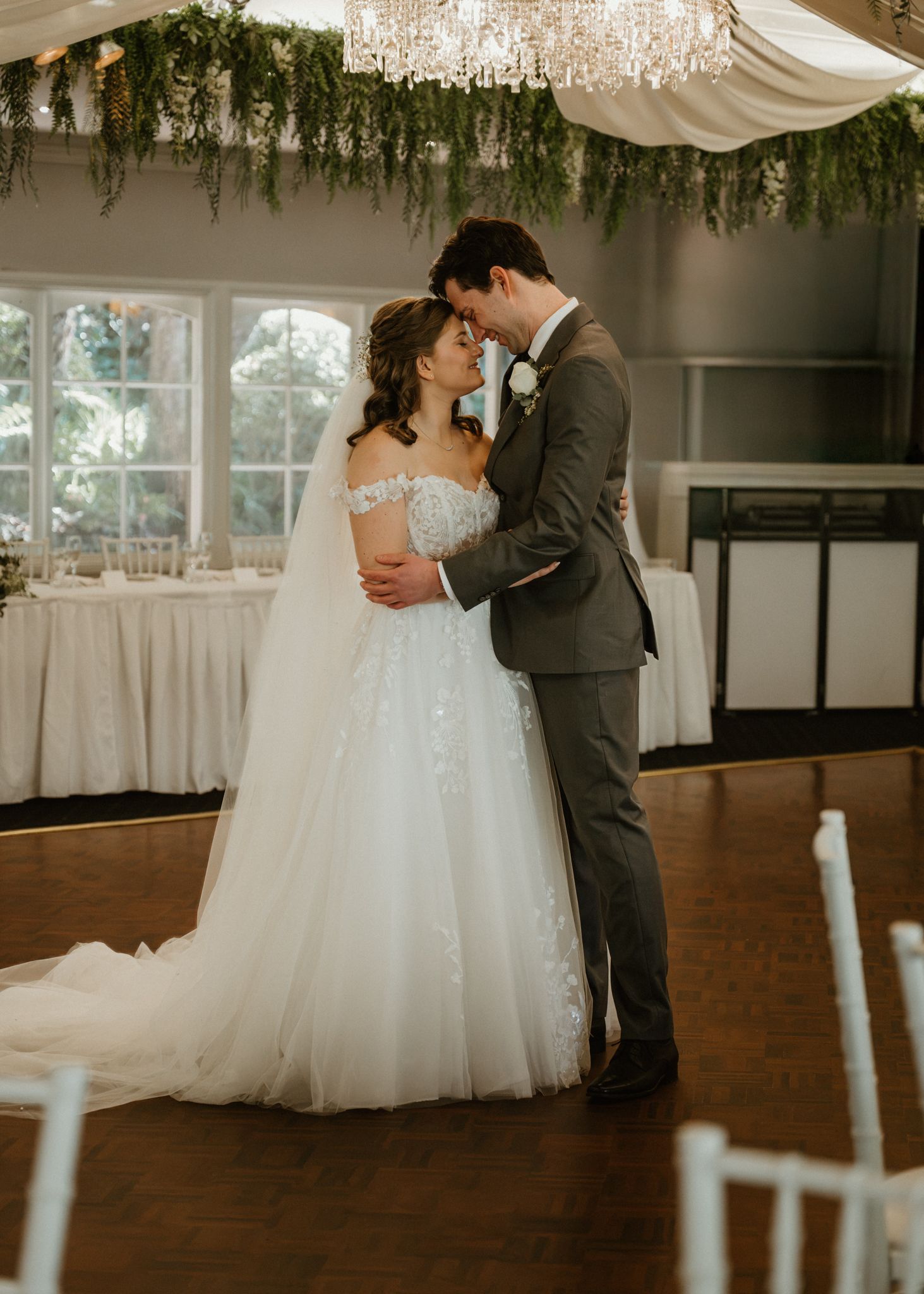 Bride in lace off-shoulder gown and groom in gray suit share romantic first dance in elegant floral reception venue.