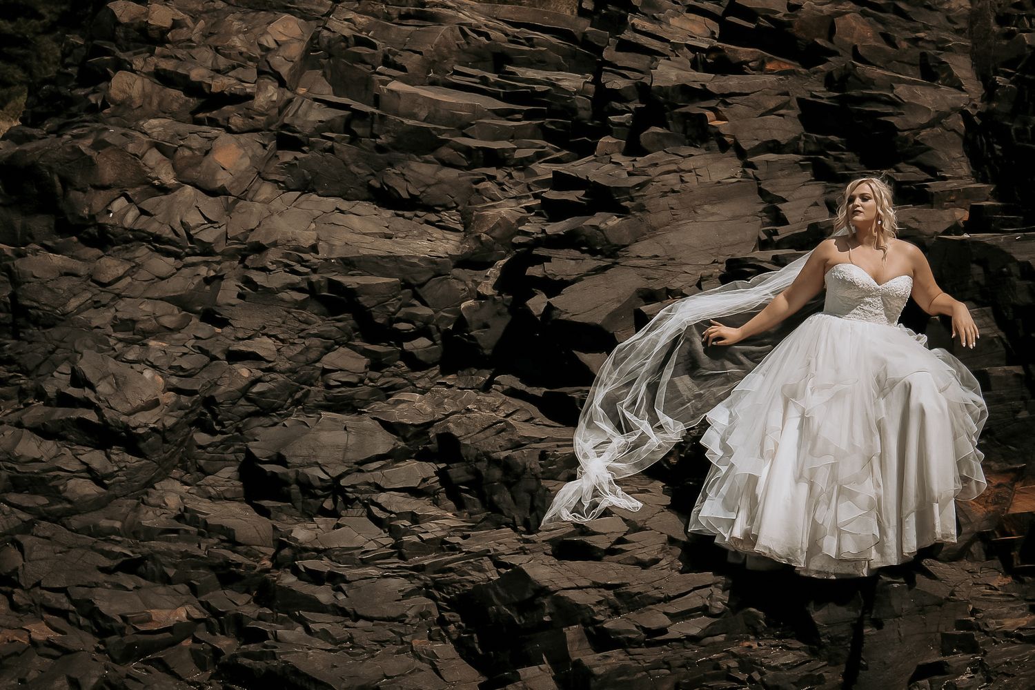 Artistic bridal portrait with flowing white dress against dark rocky backdrop.