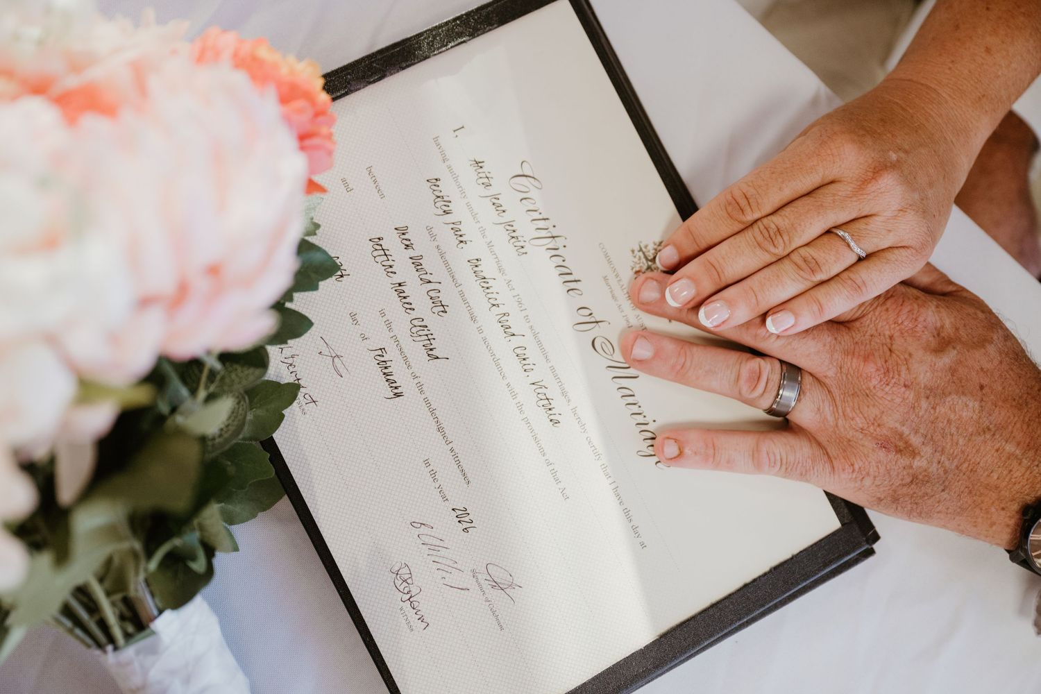 A hand with a wedding ring signs an elegant wedding guest book next to pink roses and a fountain pen.