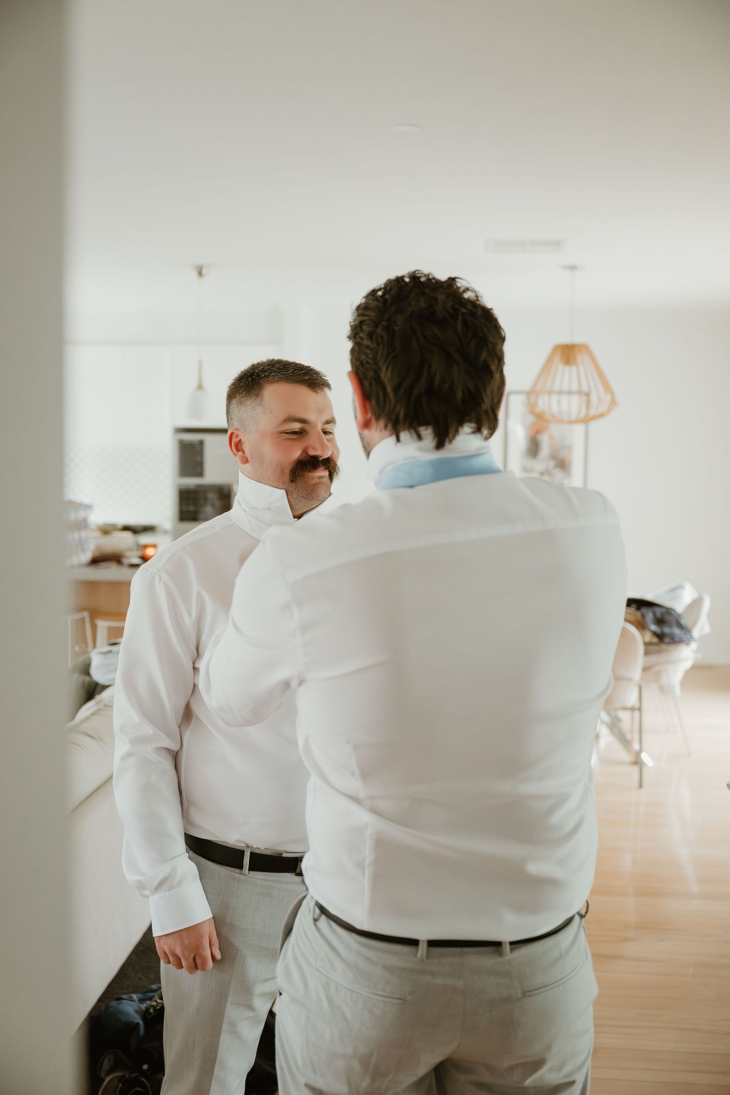 Two men in white dress shirts and gray pants share a lighthearted moment while getting ready for a wedding.