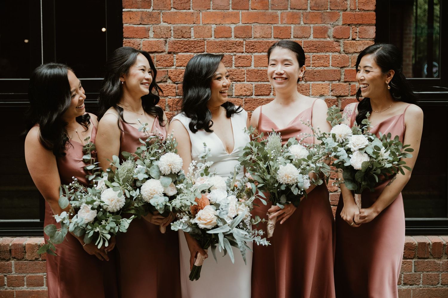 Wedding party members in burgundy and pink dresses share a candid moment while holding white floral bouquets.