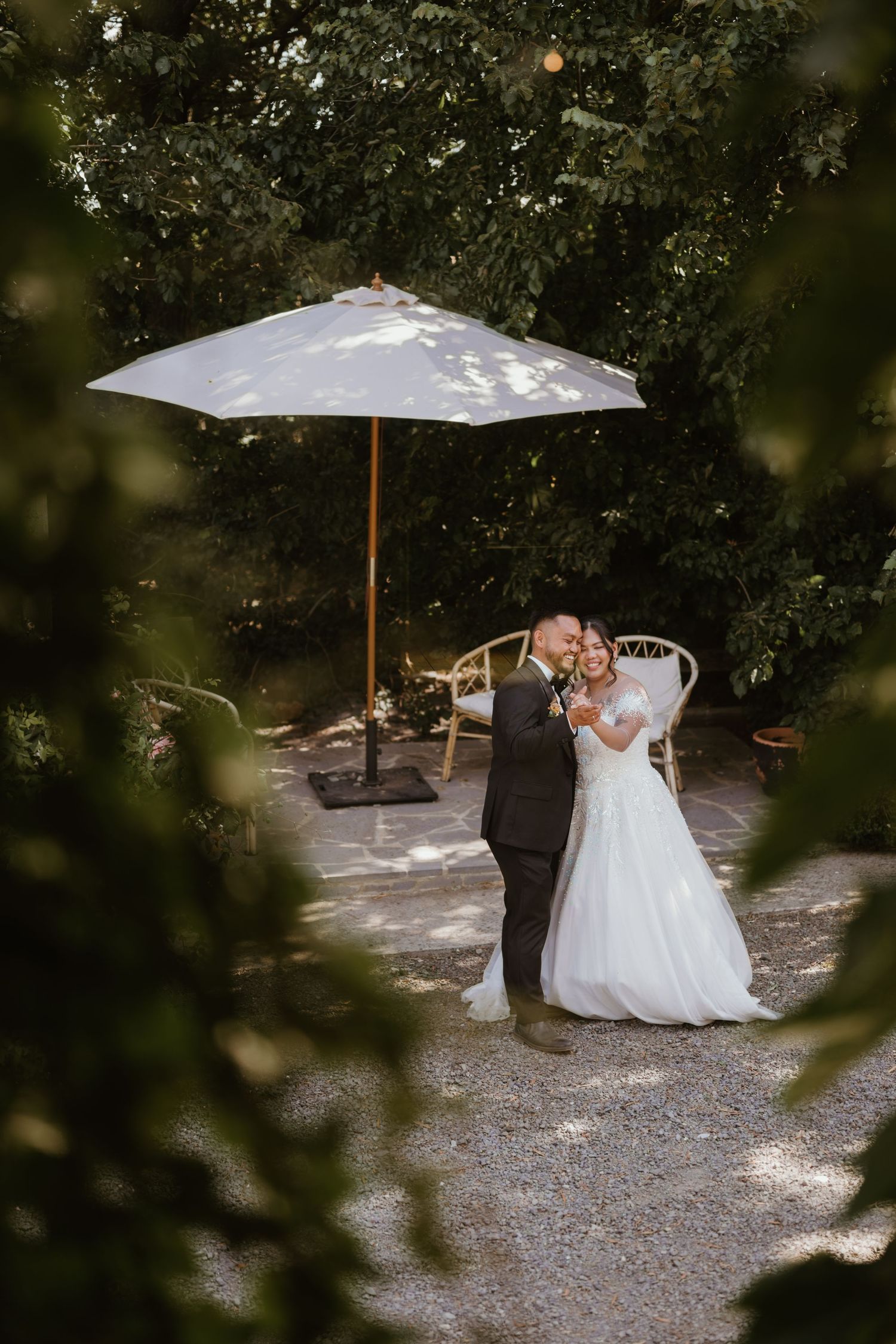 A couple shares an intimate moment under a white umbrella in a garden setting during their wedding photoshoot.
