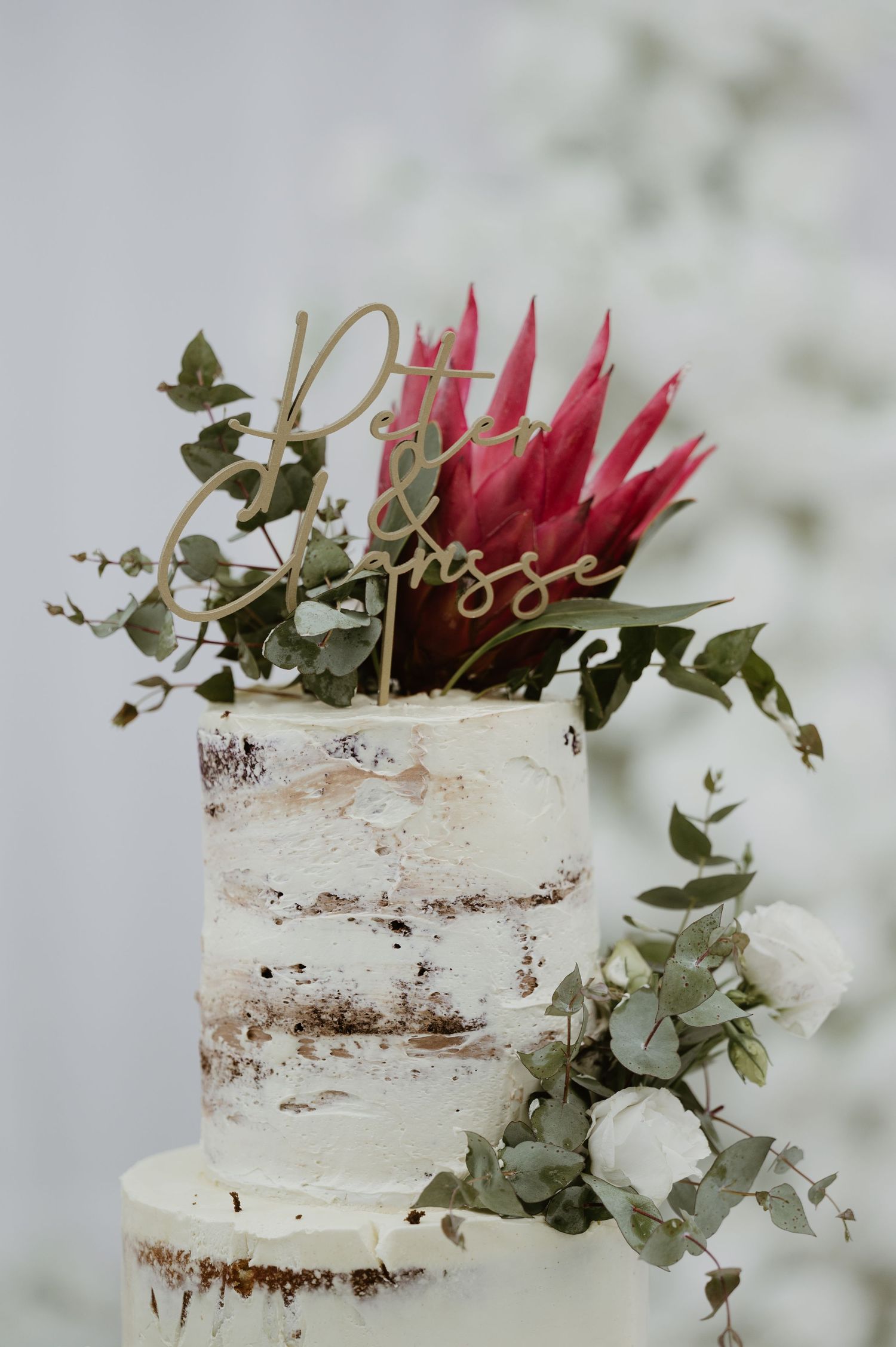 Semi-naked white wedding cake decorated with eucalyptus leaves and a striking red protea flower on top.