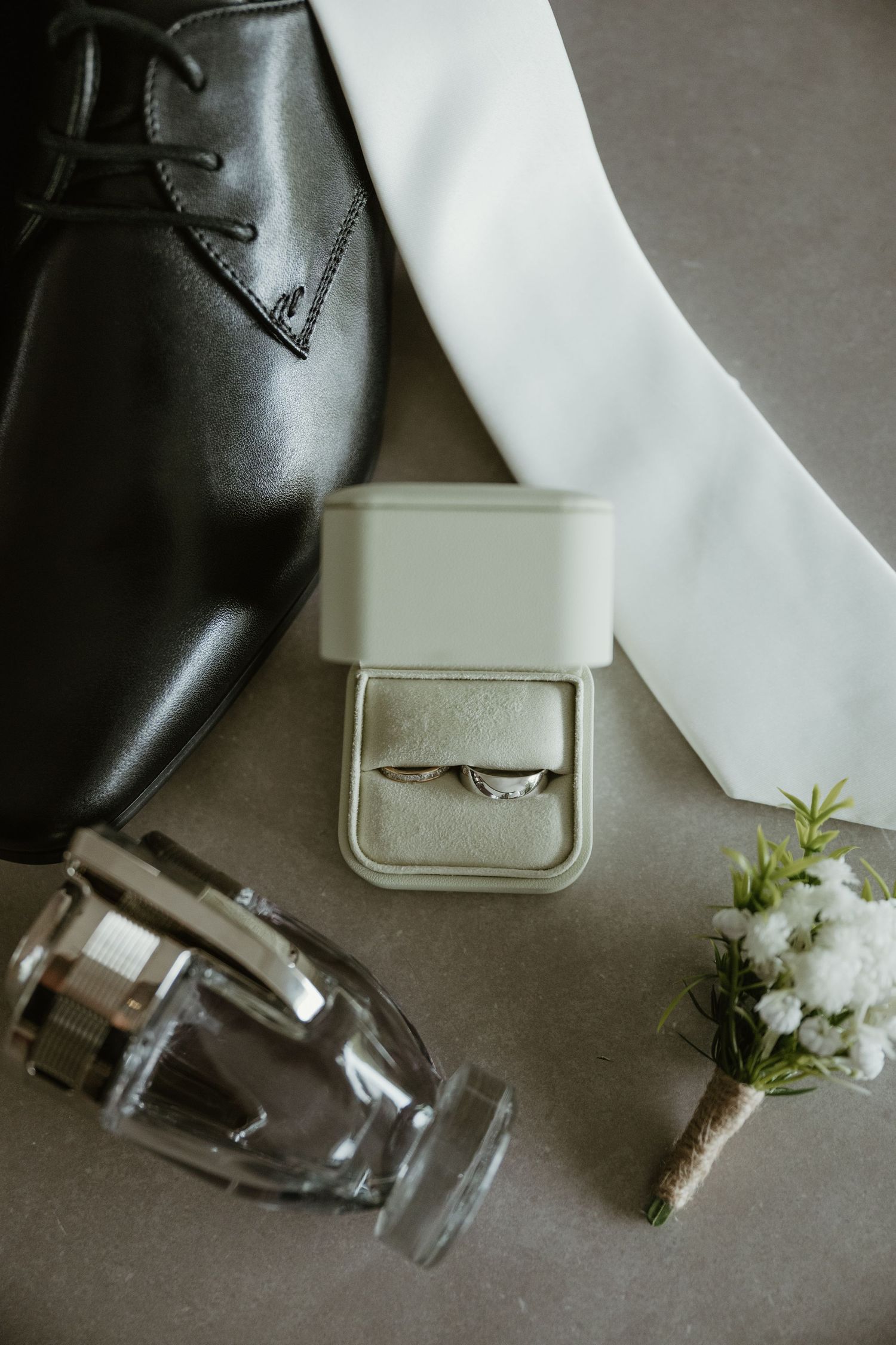Groom wedding details flat lay with ring box, white tie, boutonniere, cologne bottle, and black dress shoe.
