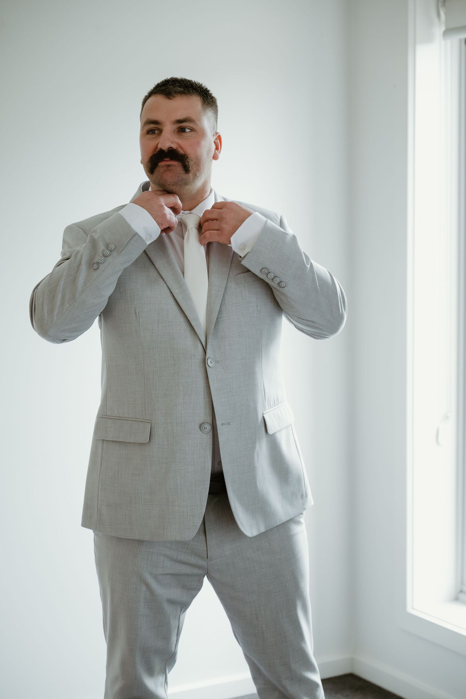 A man in a light gray suit adjusts his bow tie while standing near a bright window.